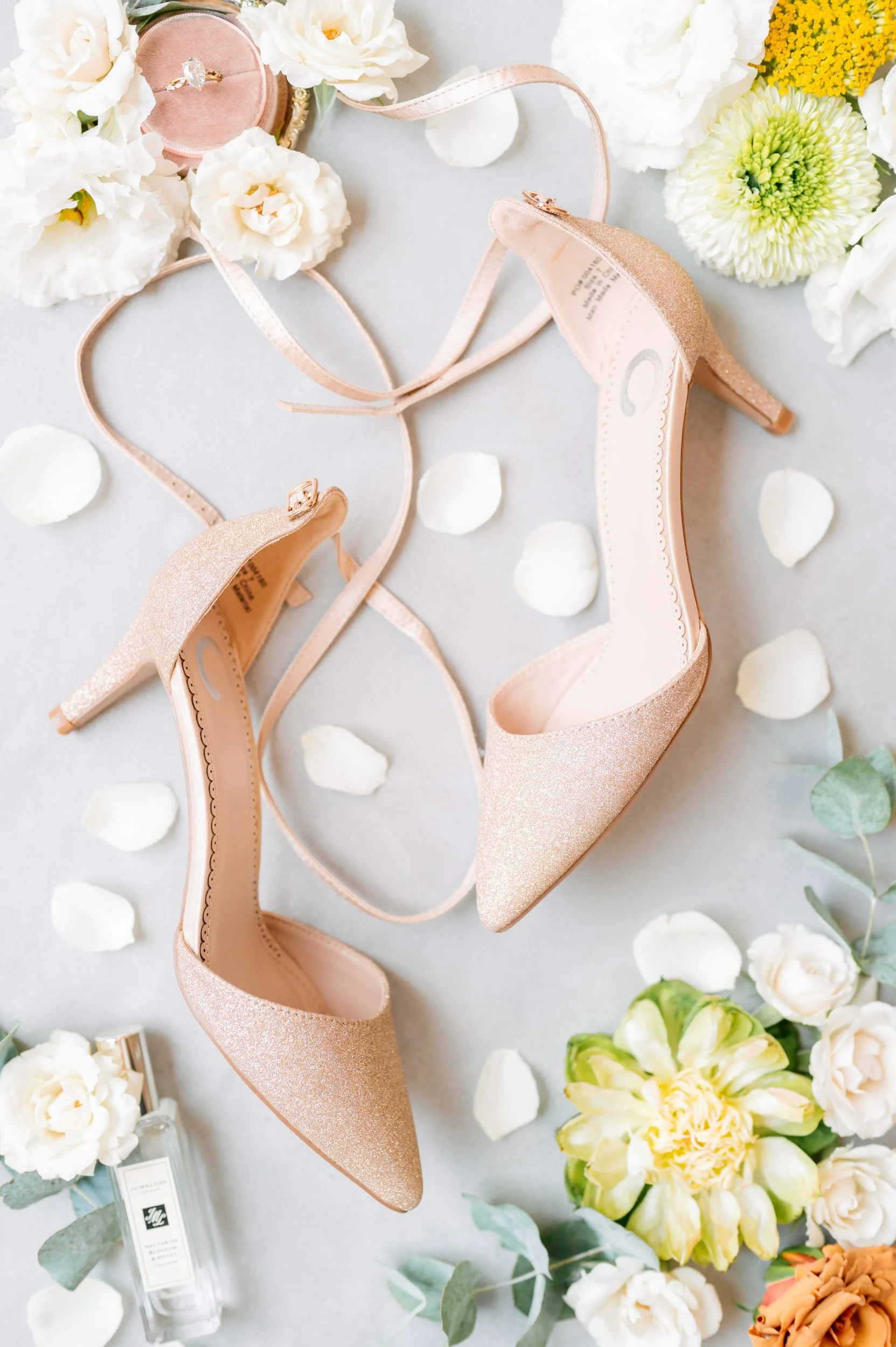 A pair of nude-colored, glittery high-heeled shoes with ankle straps surrounded by white rose petals, white and yellow flowers, and a small perfume bottle on a light surface, possibly for a wedding or special occasion.