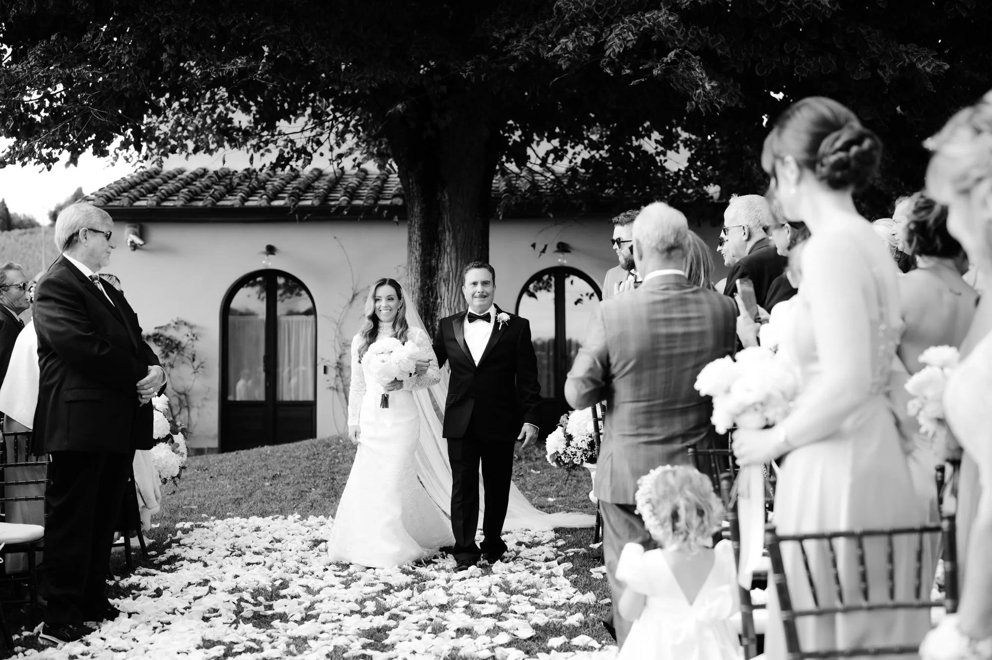 Black and white photo of a wedding ceremony outdoors, with the bride and groom walking down the aisle surrounded by guests holding bouquets, under a large tree.