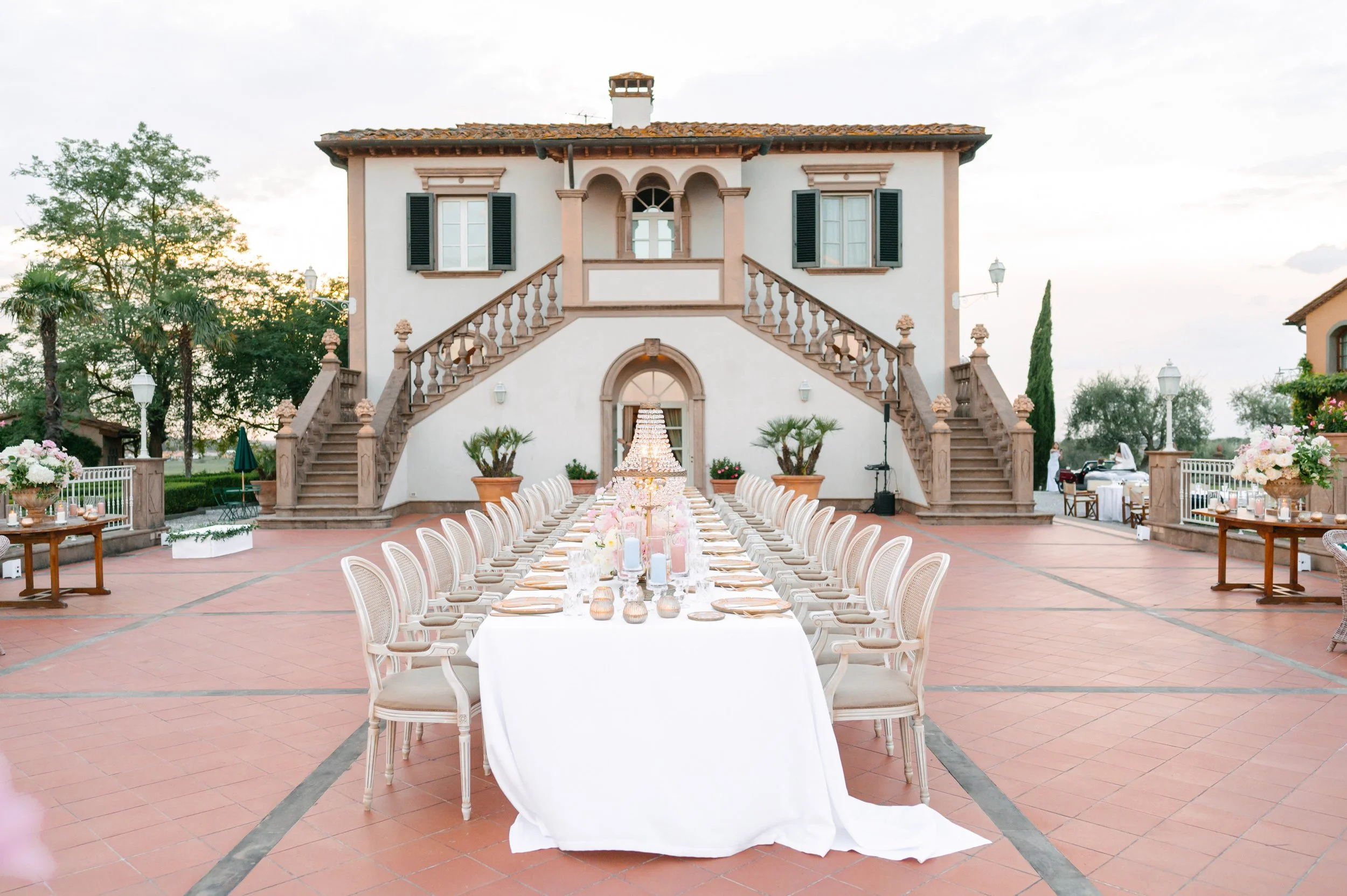 Outdoor wedding reception setup with a long white table, elegant floral centerpieces, and cream-colored chairs in front of a Mediterranean-style house with stone stairs and lush greenery.