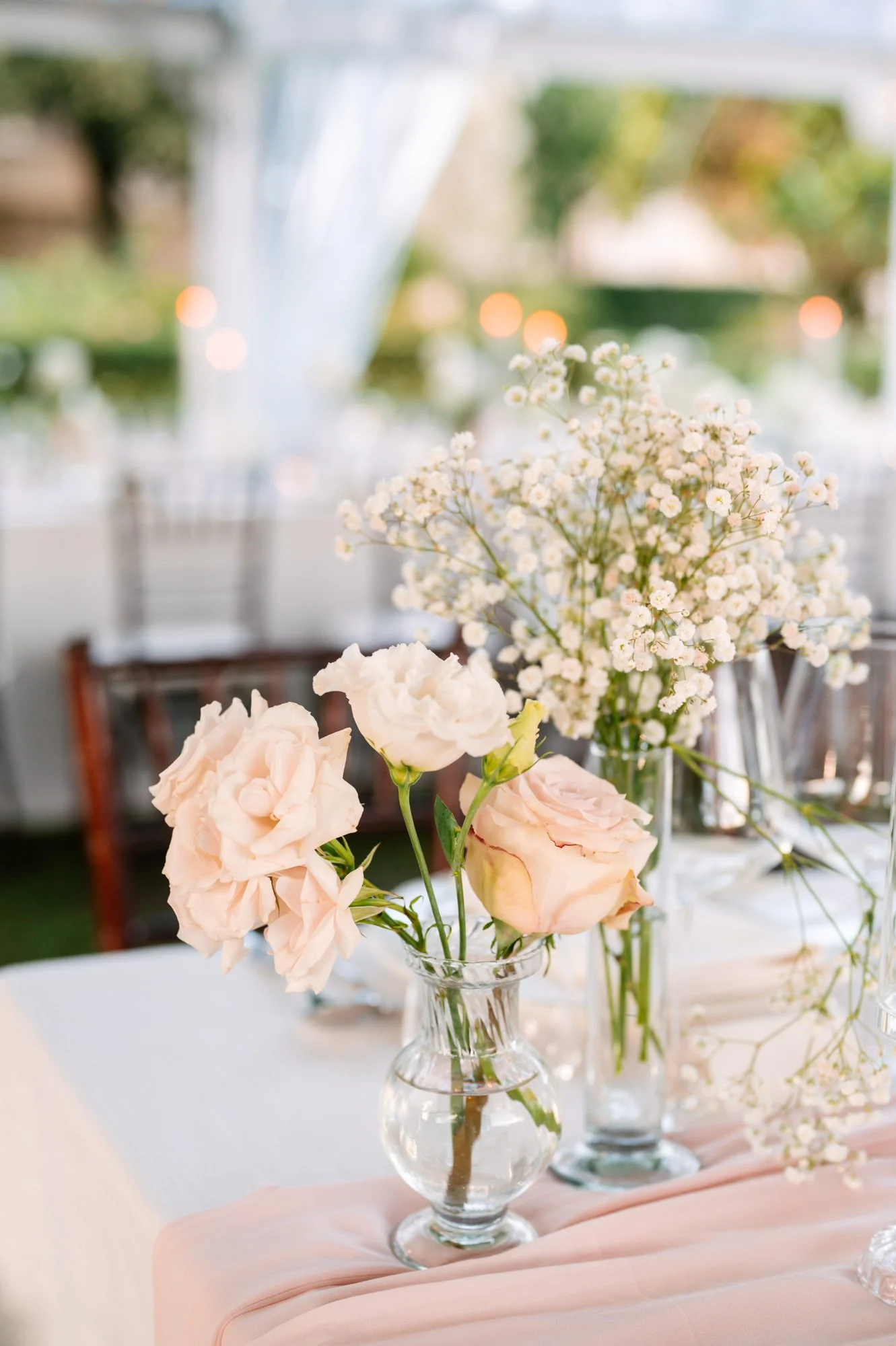 Soft pink roses and small white flowers in clear glass vases on a table with a light pink cloth, outdoors with blurred greenery and bokeh lights in the background.