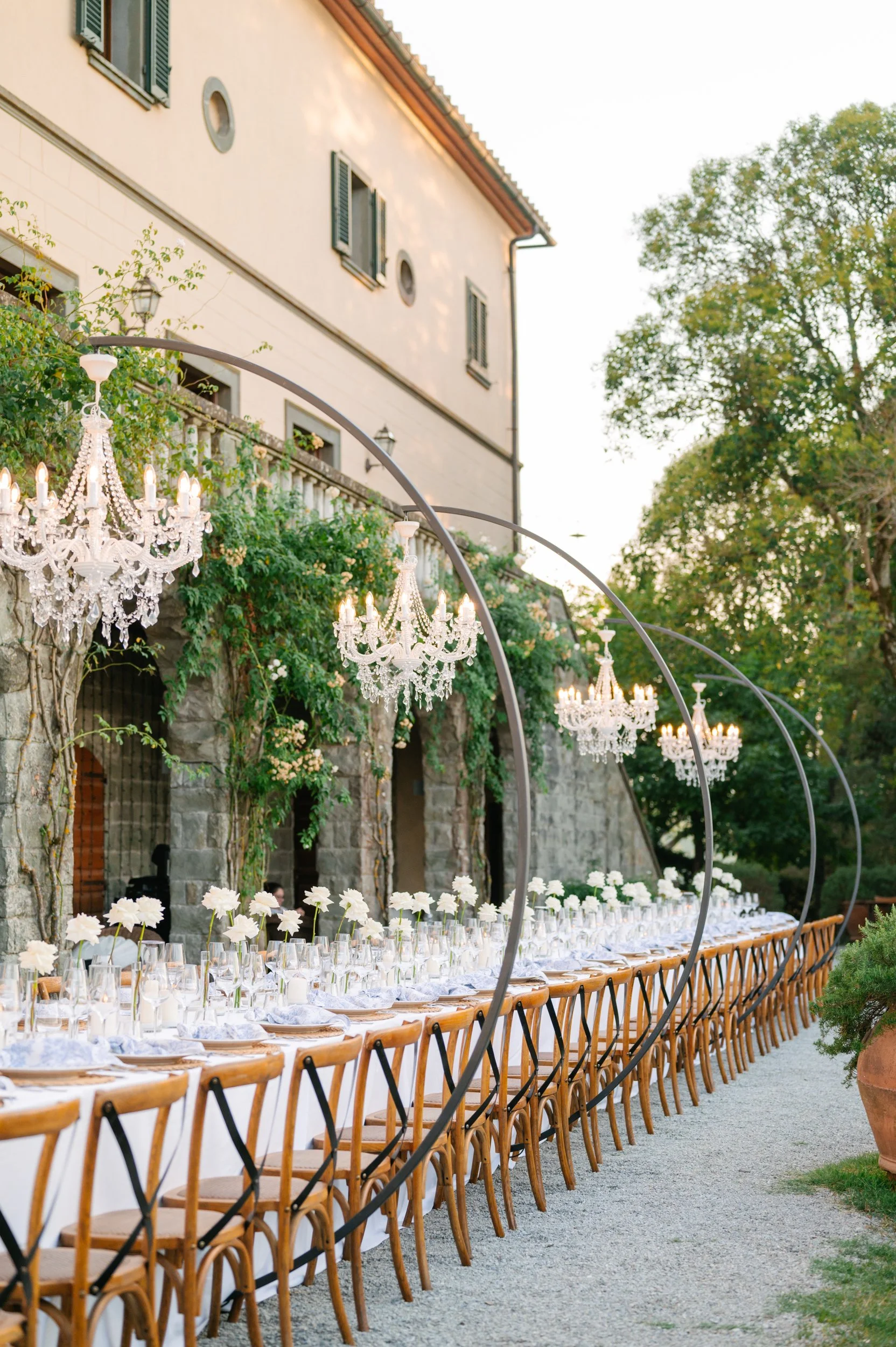 Long outdoor dining table set with white tablecloths, white flowers in glass vases, and glassware, under hanging chandeliers, with a stone building and greenery in the background.