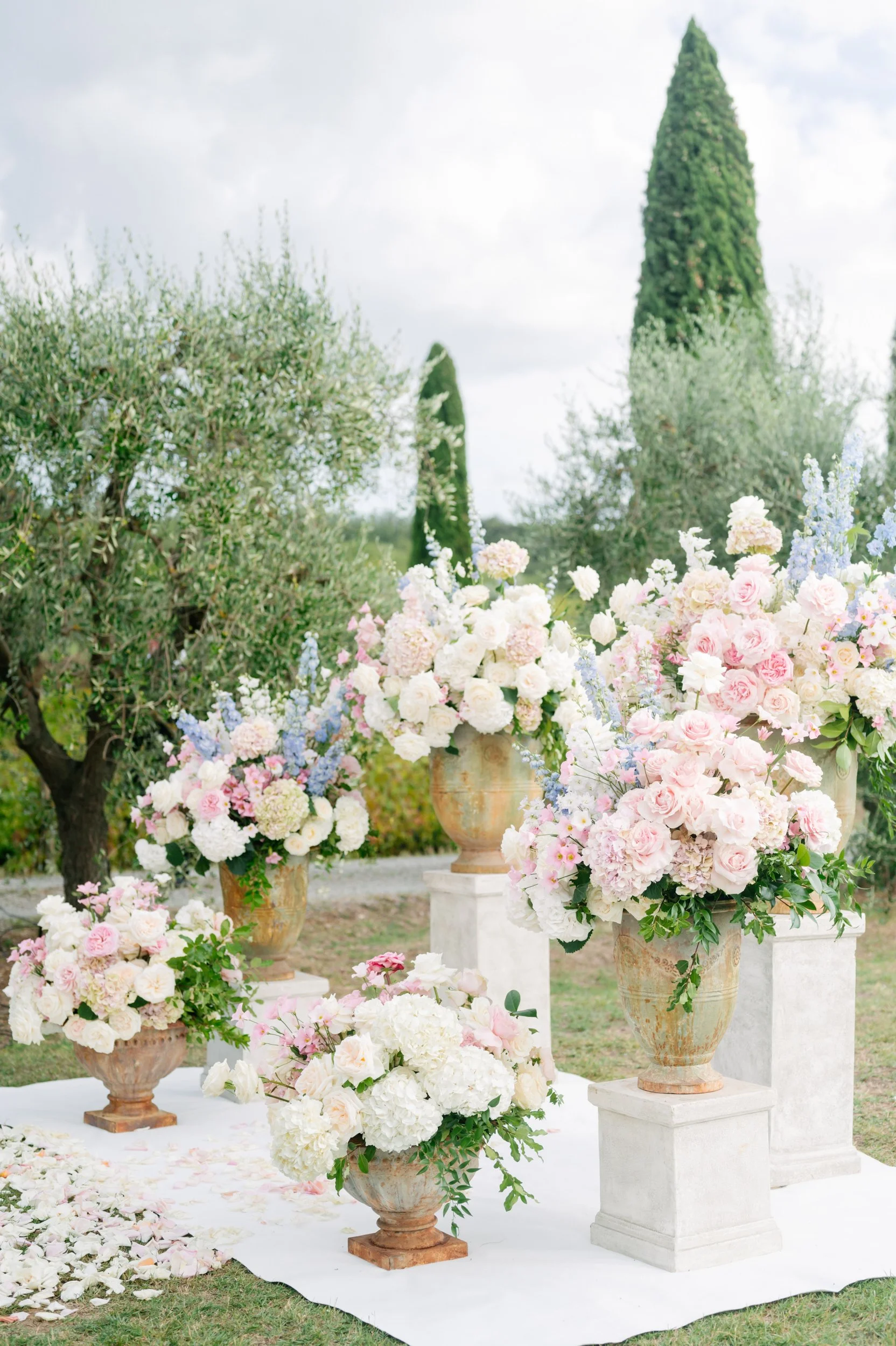 A floral arrangement display with white, pink, and pastel-colored flowers in large urns and vases, set outdoors on a white cloth, with trees and a cloudy sky in the background.