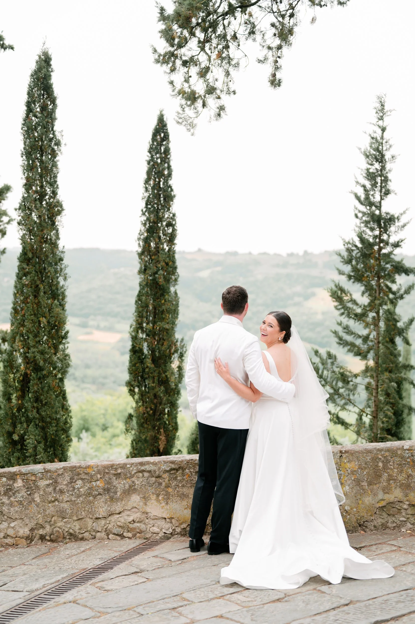 A bride and groom stand on a stone terrace, facing each other and smiling, with a scenic landscape of green trees and rolling hills in the background.