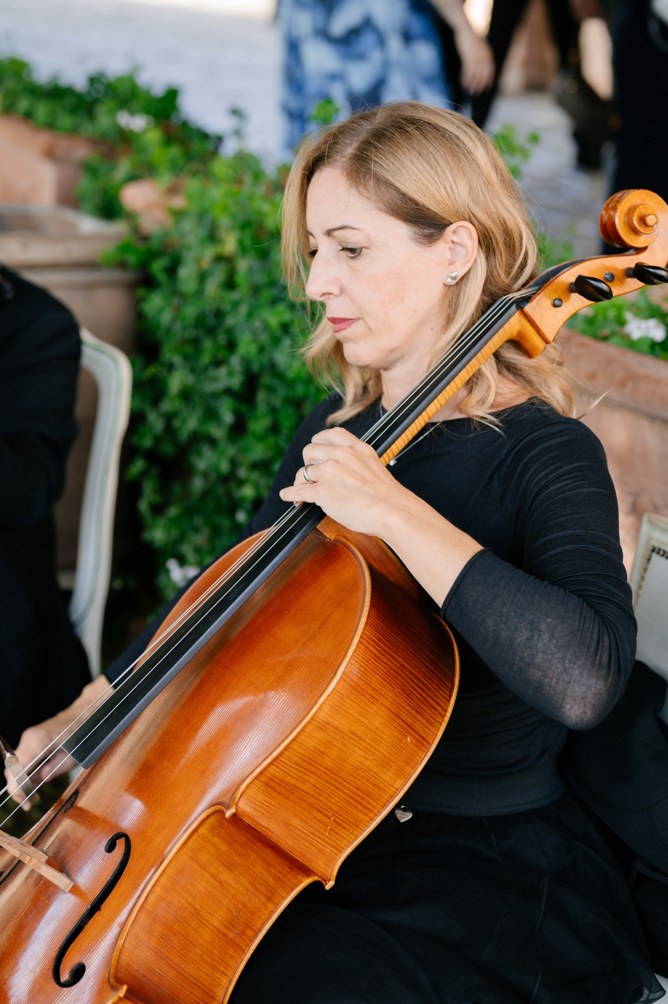 A woman with blonde hair playing a cello outdoors, dressed in a black top, with greenery and people in the background.