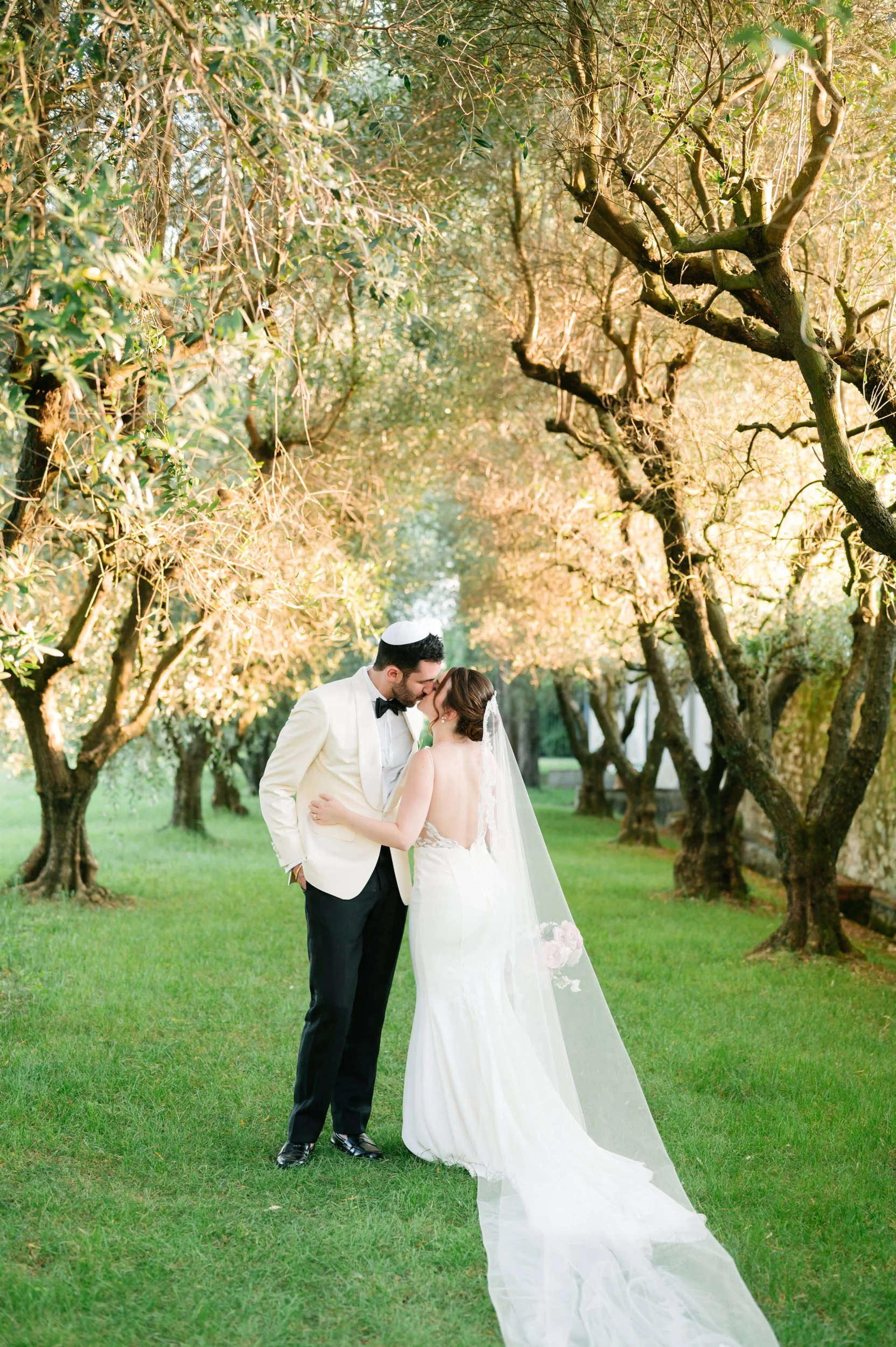 A bride and groom kissing in a lush, green outdoor setting with trees and sunlight.