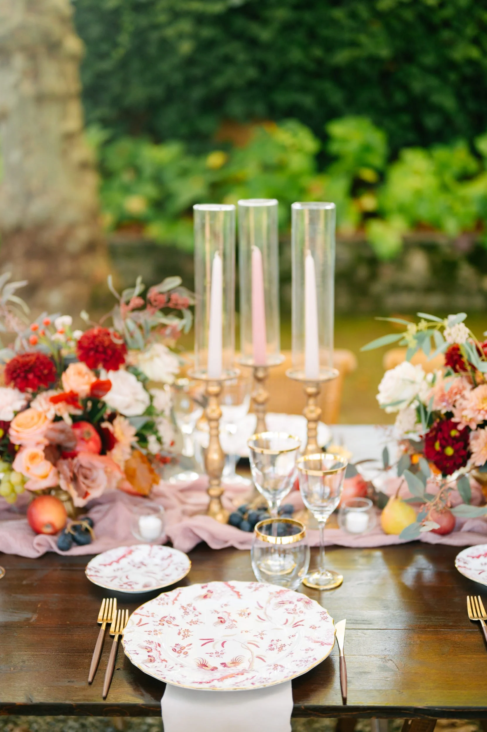 Elegant outdoor table setting with floral centerpiece, gold-rimmed glasses, taper candles in gold candlesticks, and patterned dinnerware amid lush greenery.