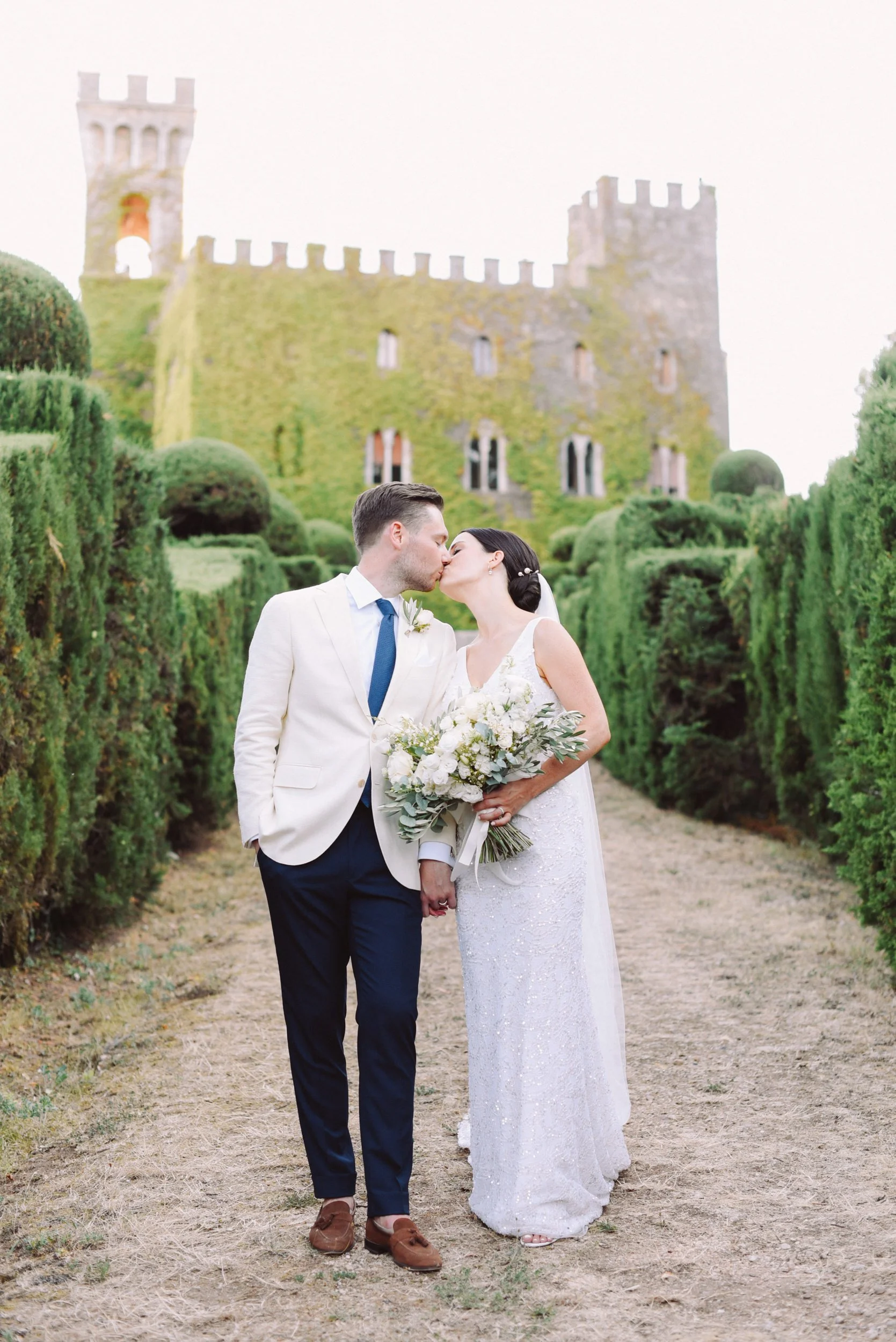 A bride and groom kiss on a dirt path, holding hands, in front of a historic castle with green moss and ivy, surrounded by lush green hedges.