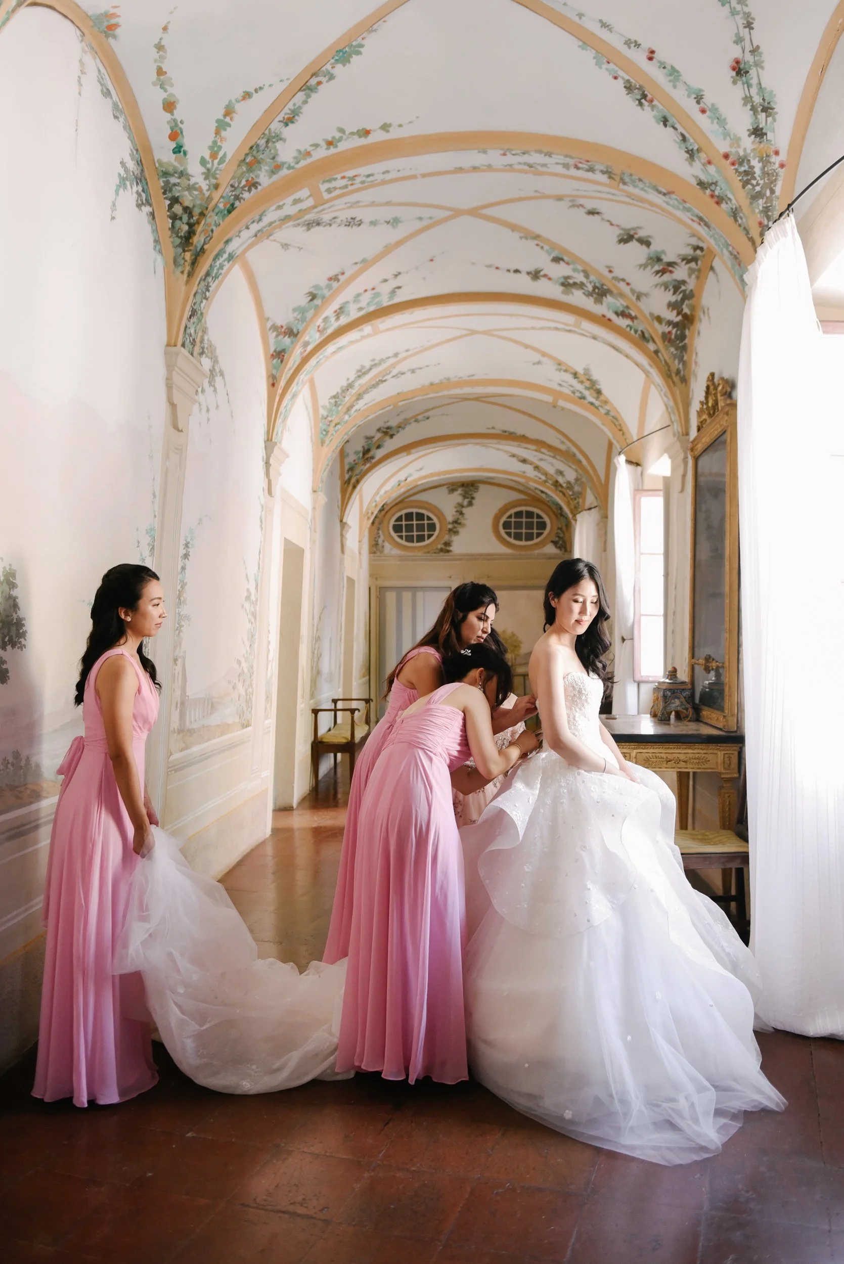 A bride in a white wedding gown is getting ready in a decorated room with four women in pink dresses helping her.
