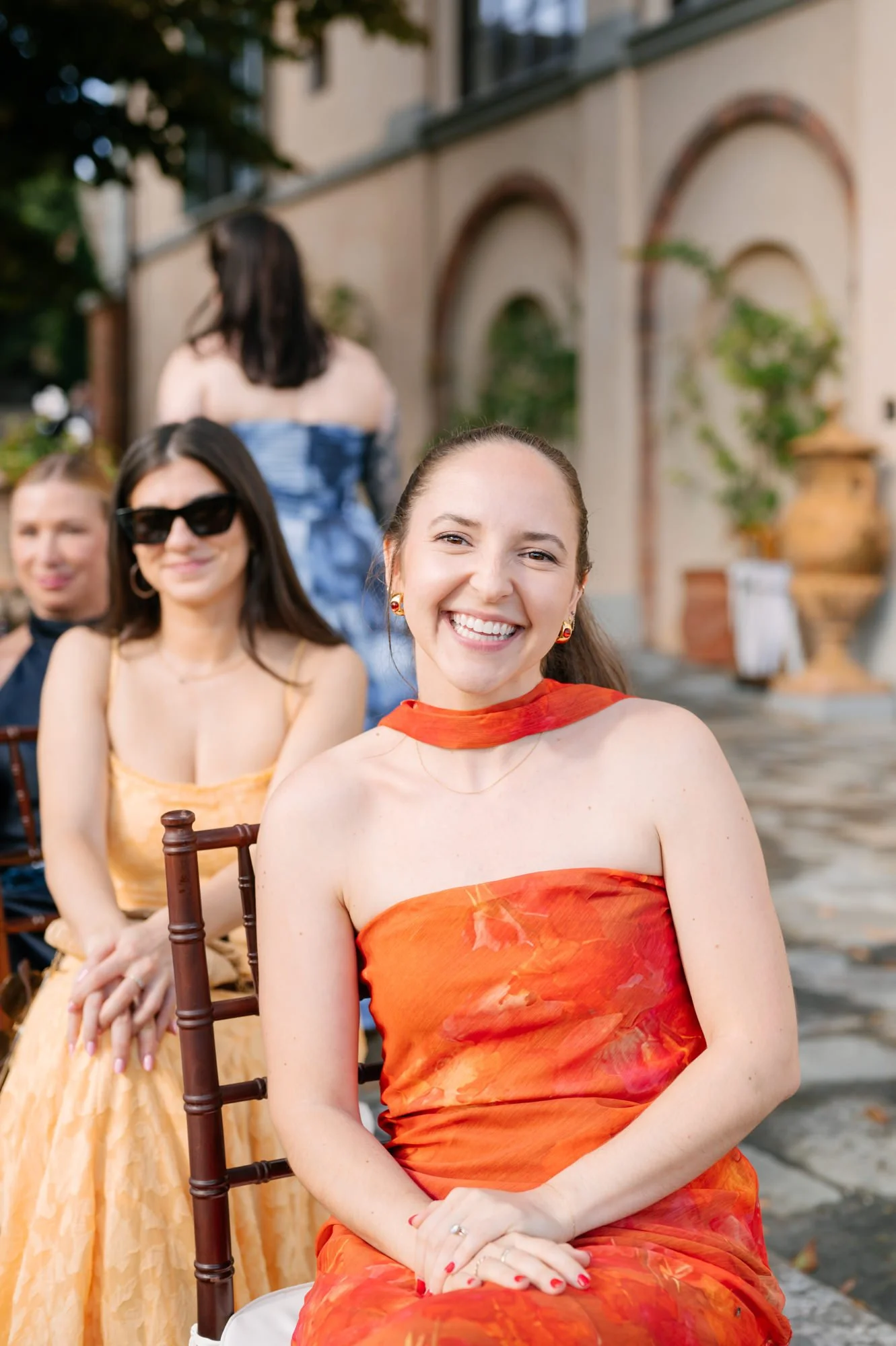 A group of women dressed in colorful dresses sitting outdoors, smiling, with a building and greenery in the background.