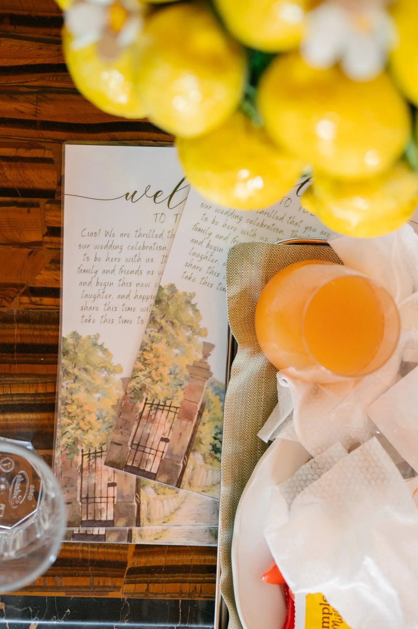A wedding invitation with handwriting and a painted illustration of a gate and trees, placed on a wooden table next to a glass of orange juice and a plate with napkins.