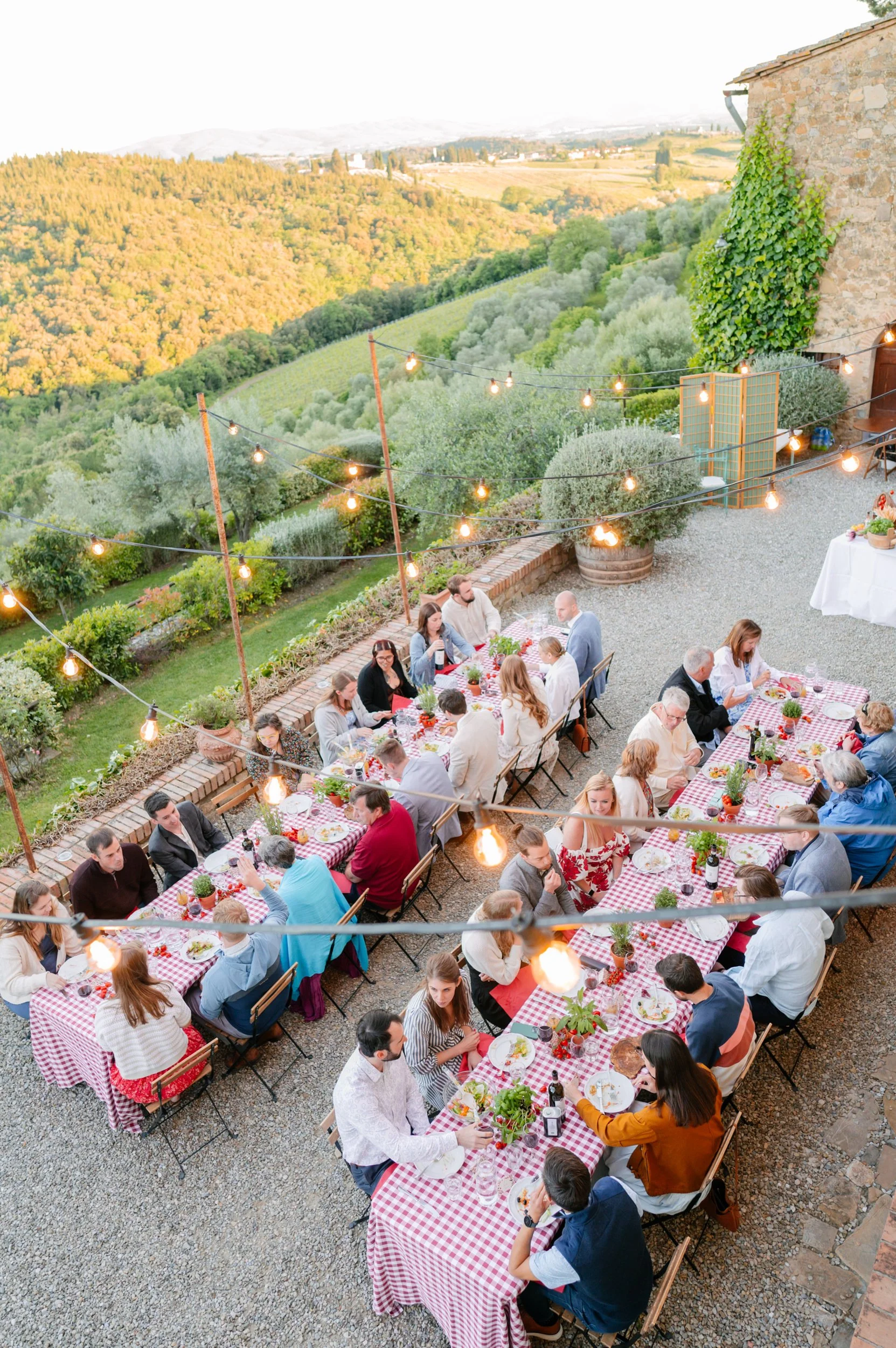 Outdoor dinner party with long tables covered in red and white checkered tablecloths, set with plates, glasses, and potted plants, illuminated by string lights against a backdrop of rolling green hills and a stone building.