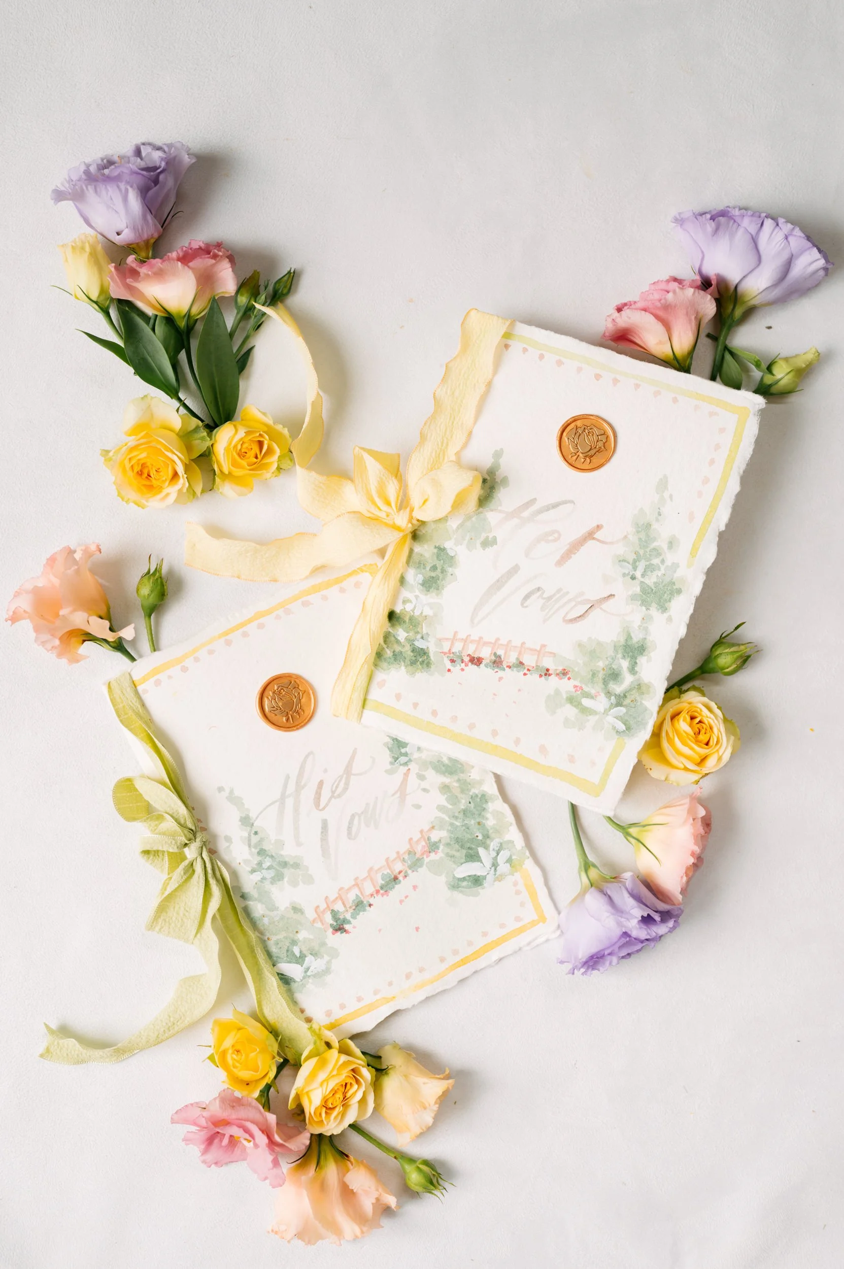 A floral wedding invitation with pastel-colored flowers attached to a yellow and green ribbon, surrounded by scattered pink, yellow, and purple flowers.