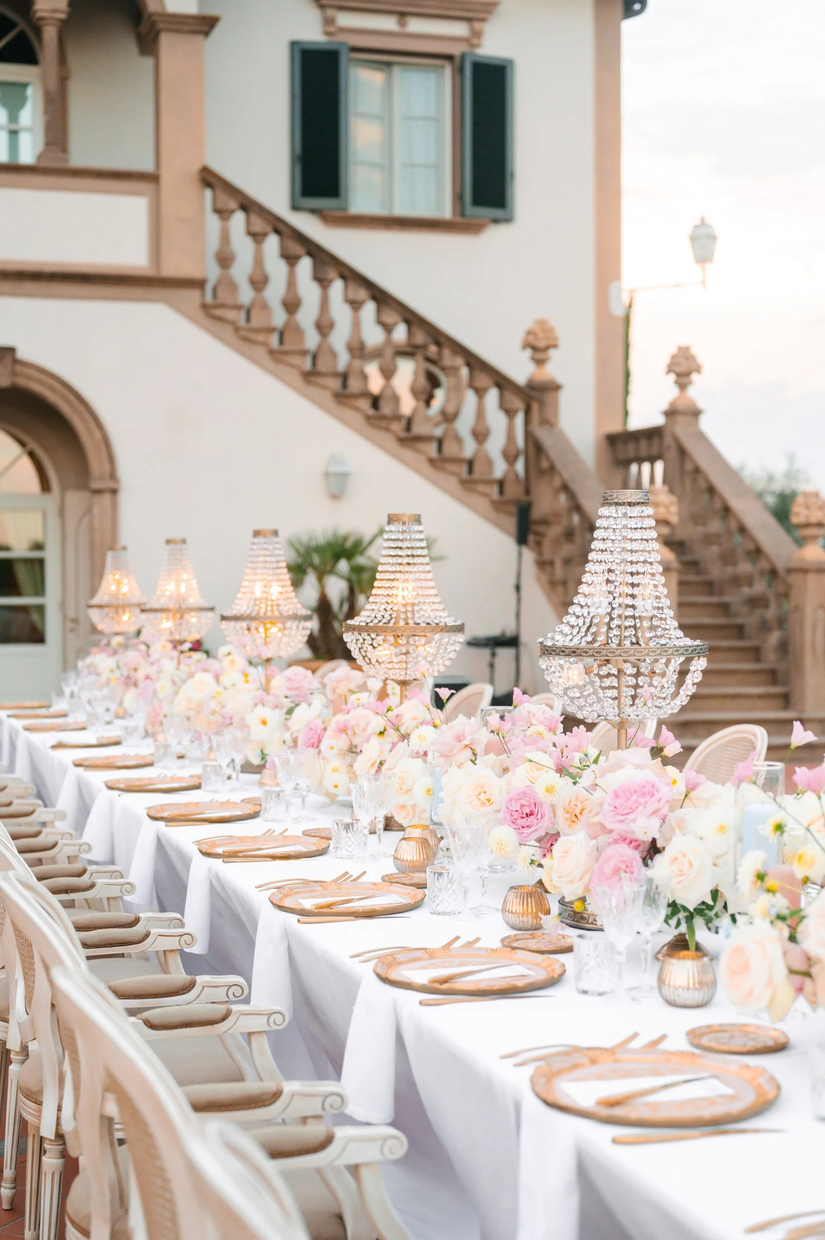Elegant outdoor banquet table set with pink and white flowers, crystal chandeliers, gold-rimmed glassware, and tan plates under a pastel-colored building with stairs and shutters.