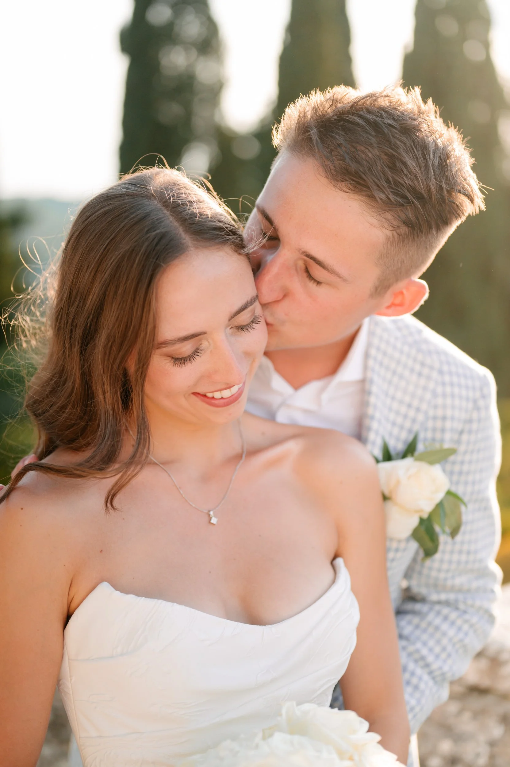 A young couple on their wedding day, with the man kissing the woman's forehead while she smiles with her eyes closed, outdoors during sunset.