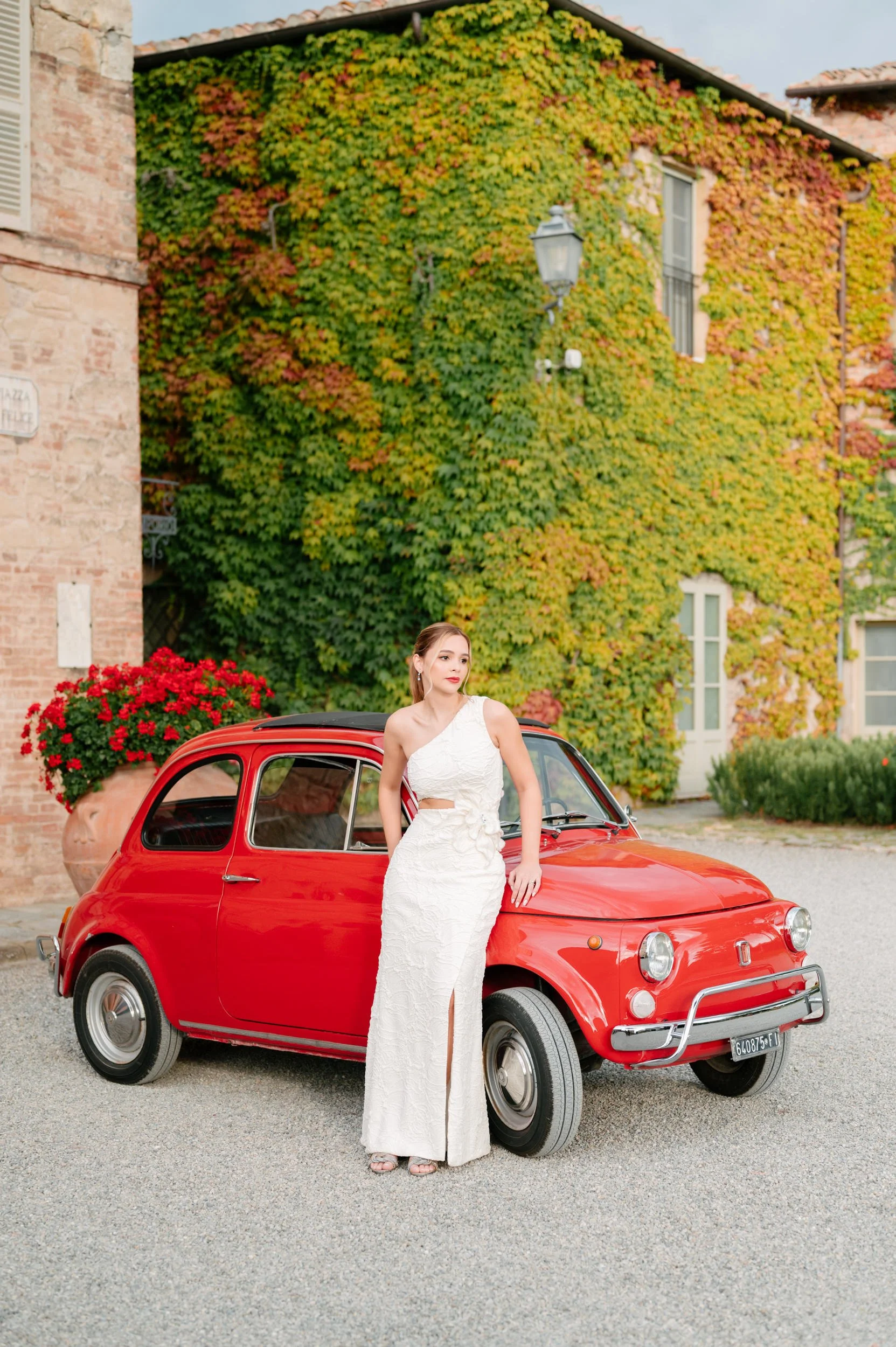 A woman in a white dress leaning against a red vintage Fiat 500 car parked on a cobblestone street, with a building covered in green and orange ivy in the background and a large red flowerpot on the sidewalk.