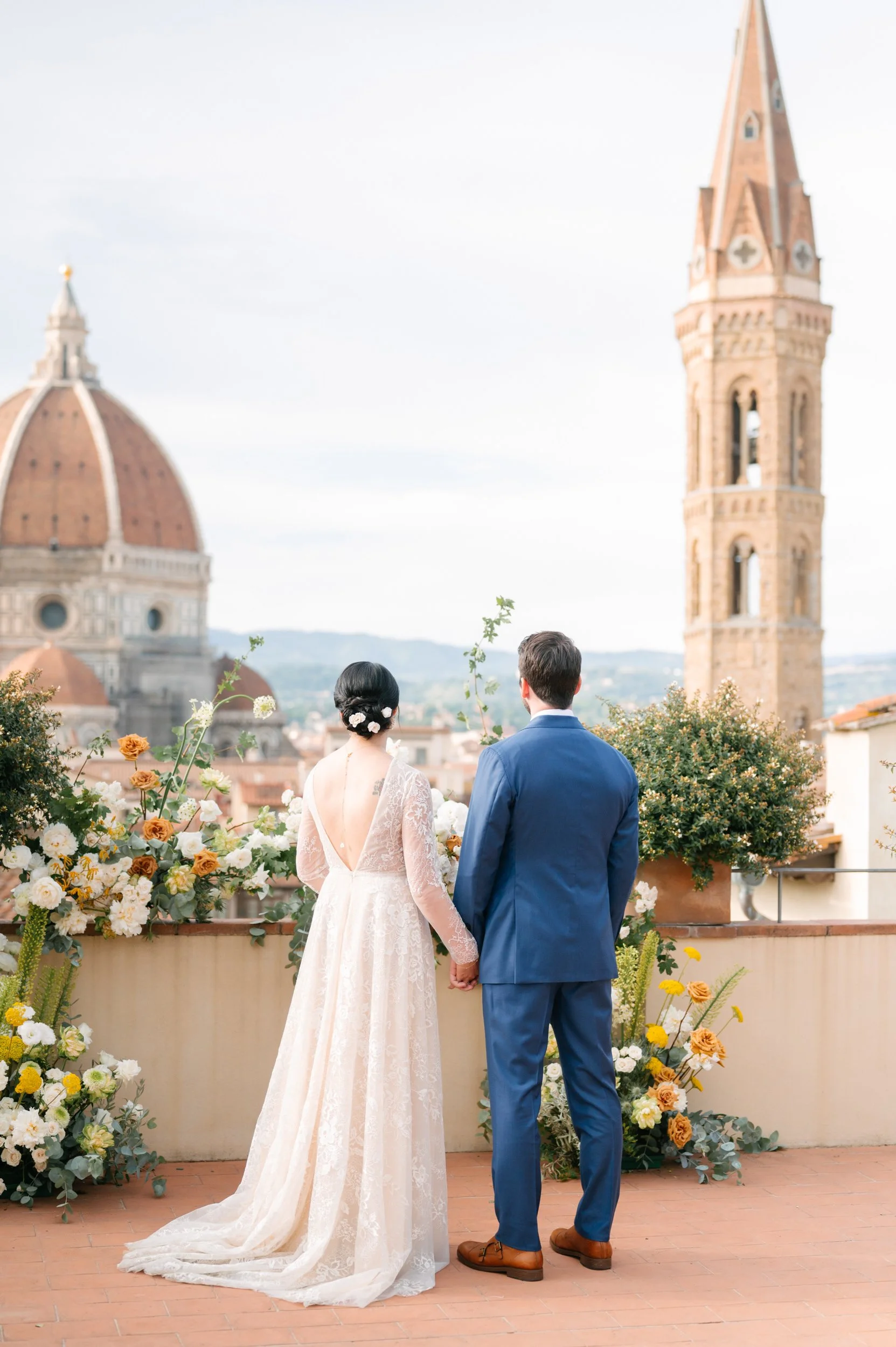A bride and groom holding hands on a balcony with floral arrangements, overlooking the Florence skyline with the Florence Cathedral and bell tower in the background.