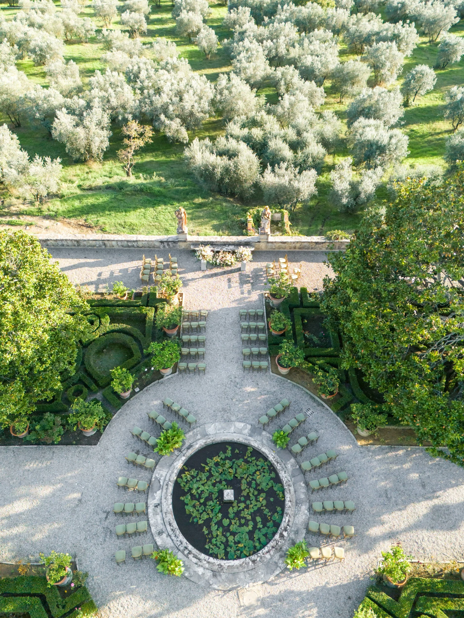 Aerial view of a lush garden with a circular pond at the center, surrounded by chairs, trees, and decorative plants, leading to a grassy area with olive trees.