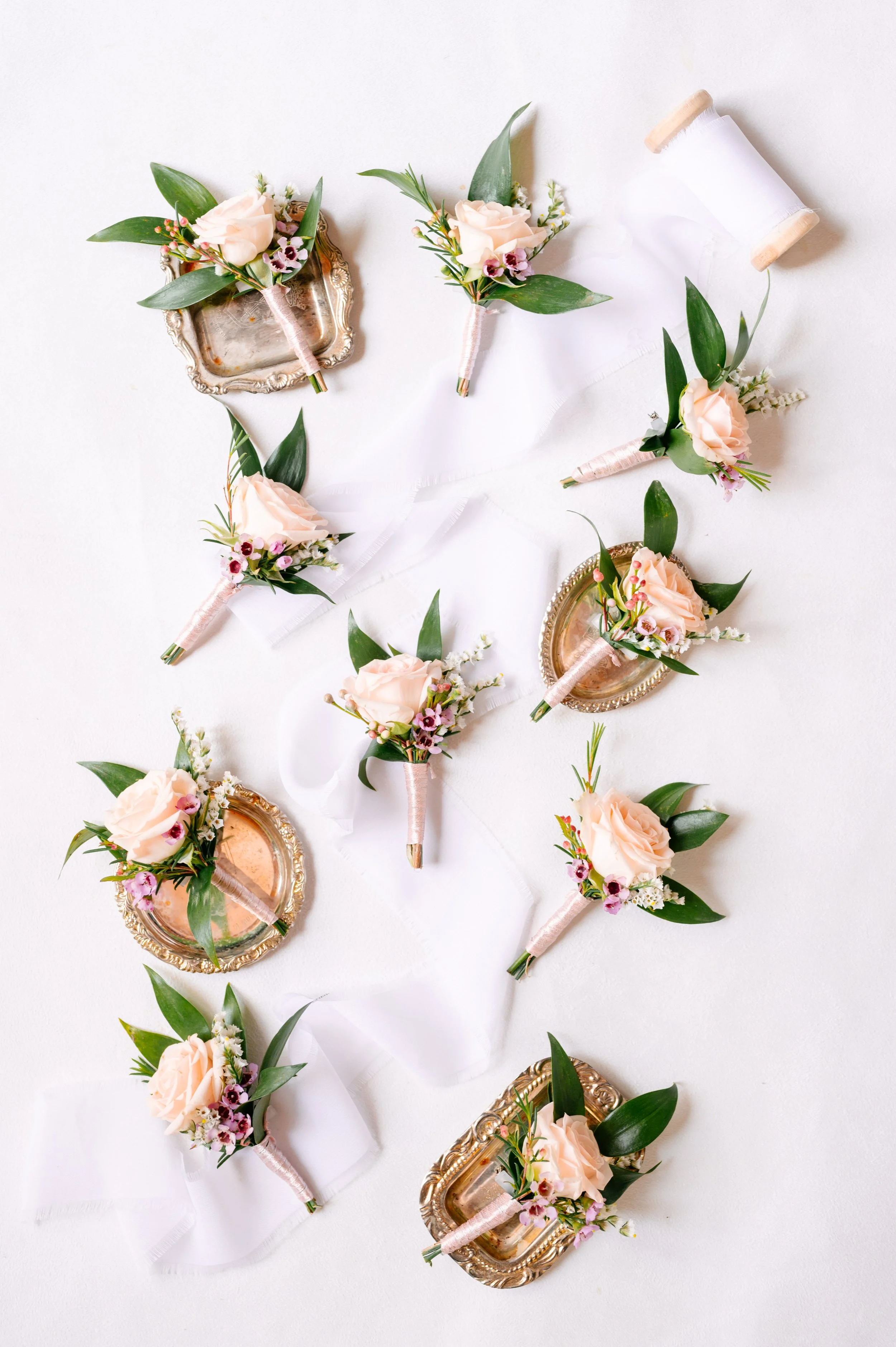 A collection of bridal boutonnieres and corsages with pale peach roses, greenery, and small pink and white flowers, arranged on a white surface, with some boutonnieres on ornate gold and silver trays.