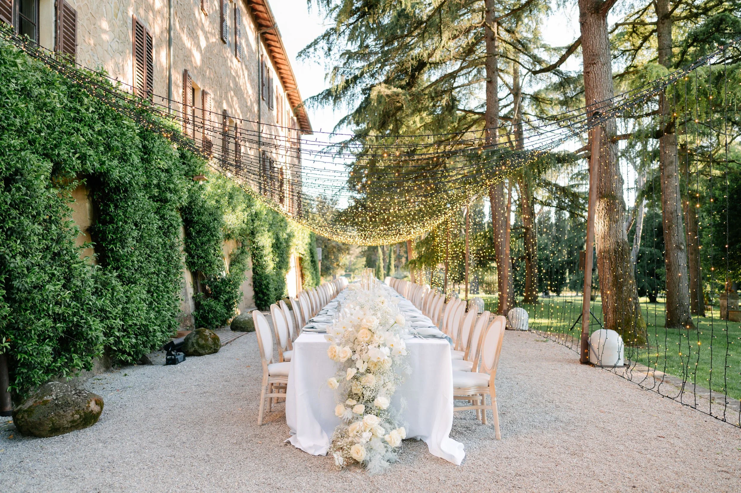 An outdoor wedding reception setup with a long table covered in a white tablecloth, adorned with white floral centerpieces. The table is surrounded by white chairs and is set under string lights hanging between trees and a building with green ivy and
