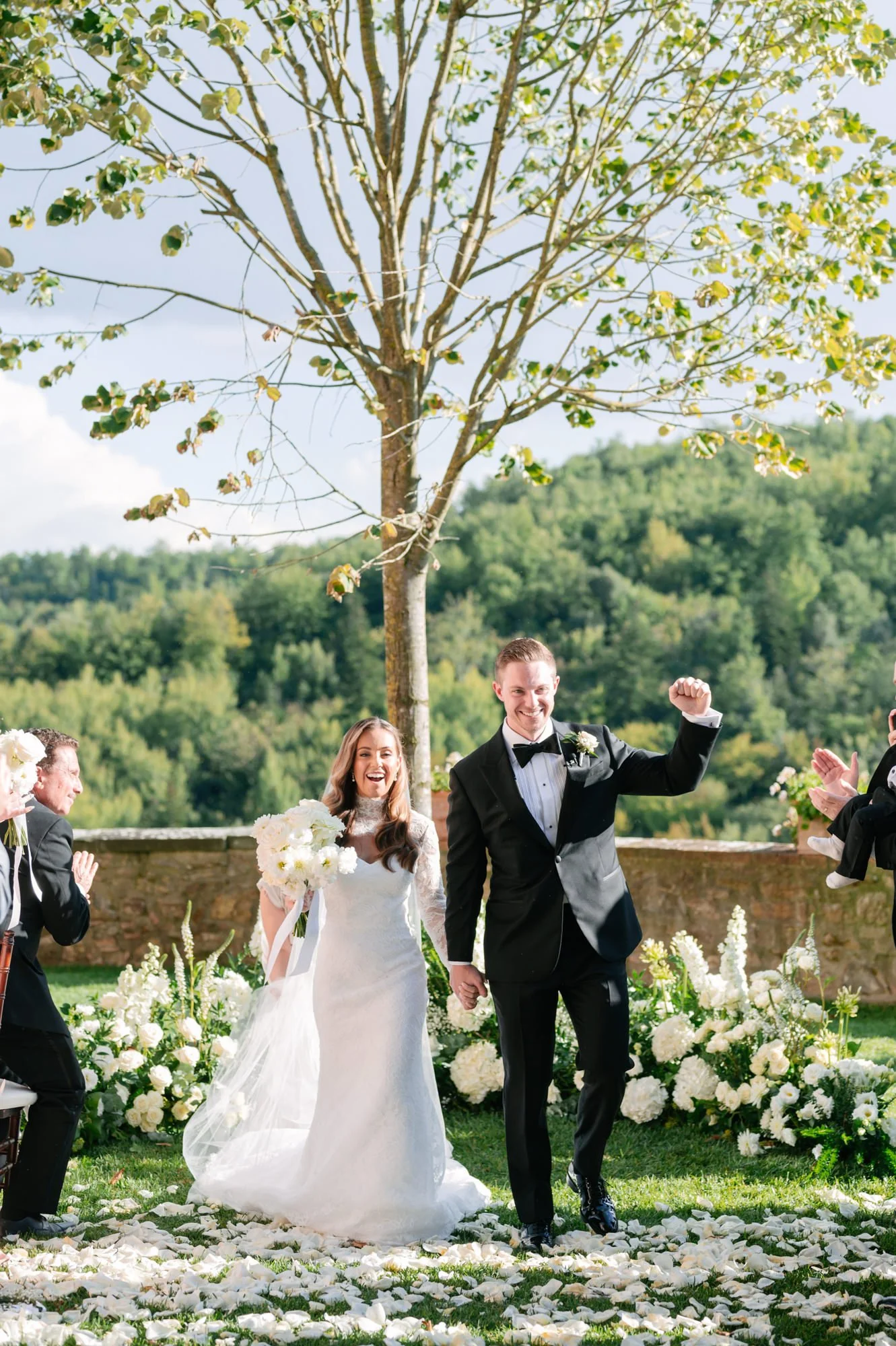 A newly married couple celebrating outdoors, with the bride holding a bouquet and the groom raising his fist in victory, surrounded by friends applauding, with flowers on the ground, a tree, and a lush green landscape in the background.