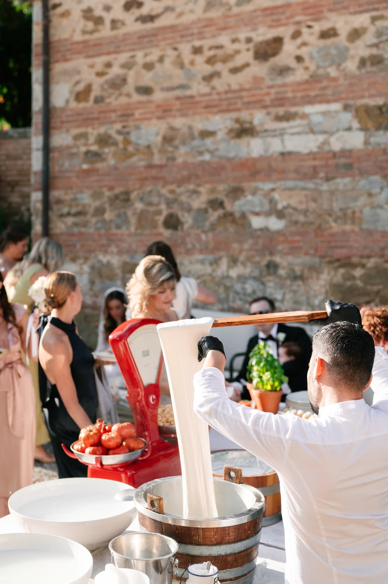 A person in white shirt and black gloves is pouring fresh mozzarella cheese from a wooden paddle into a bucket at an outdoor event with a rustic brick wall in the background. There are several women in line, some with flower hair accessories, waiting