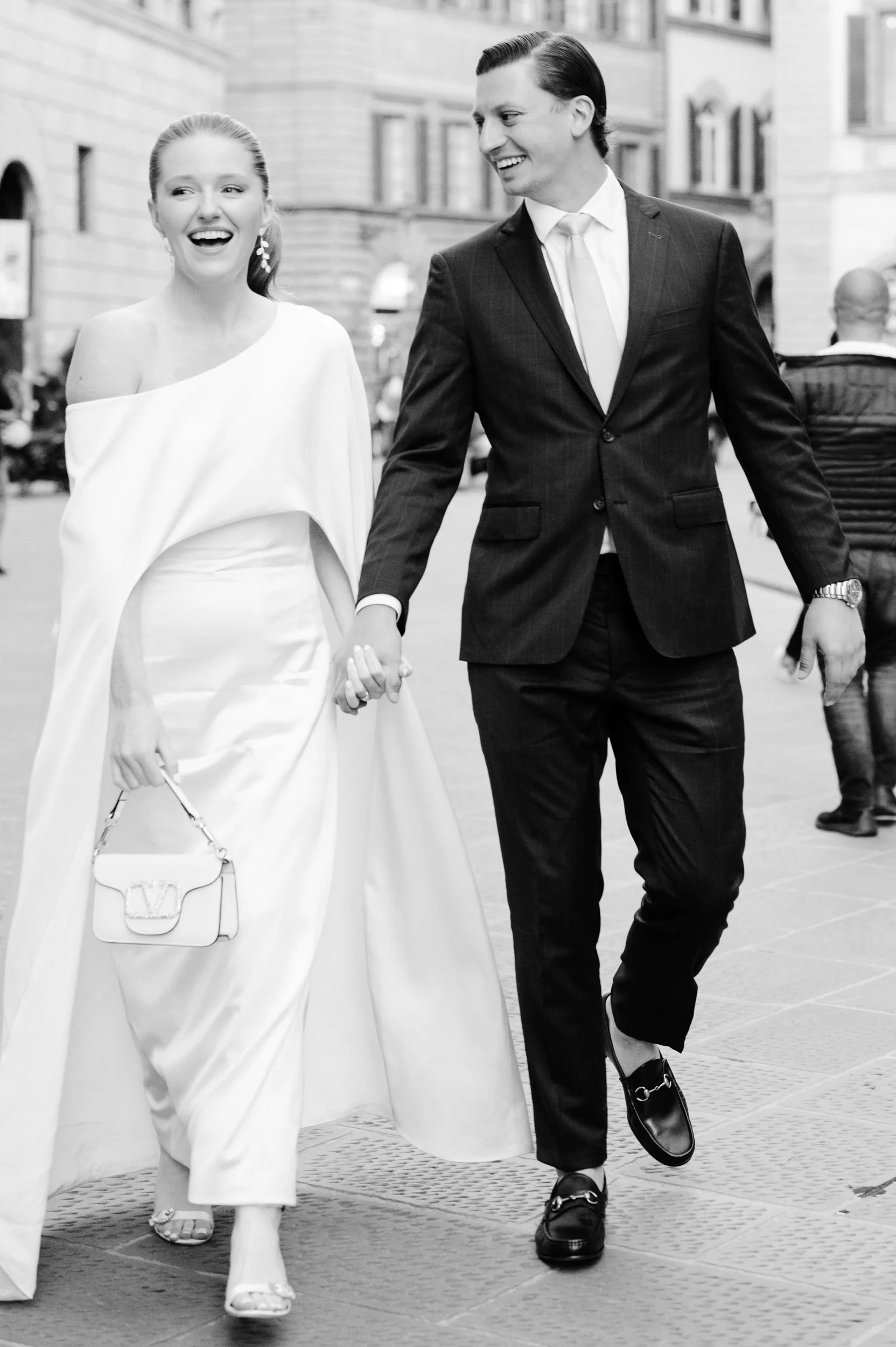 A couple walking in the city, holding hands and smiling. The woman is wearing a white dress with an asymmetrical shoulder design and carrying a small handbag. The man is dressed in a dark suit and tie, wearing loafers.