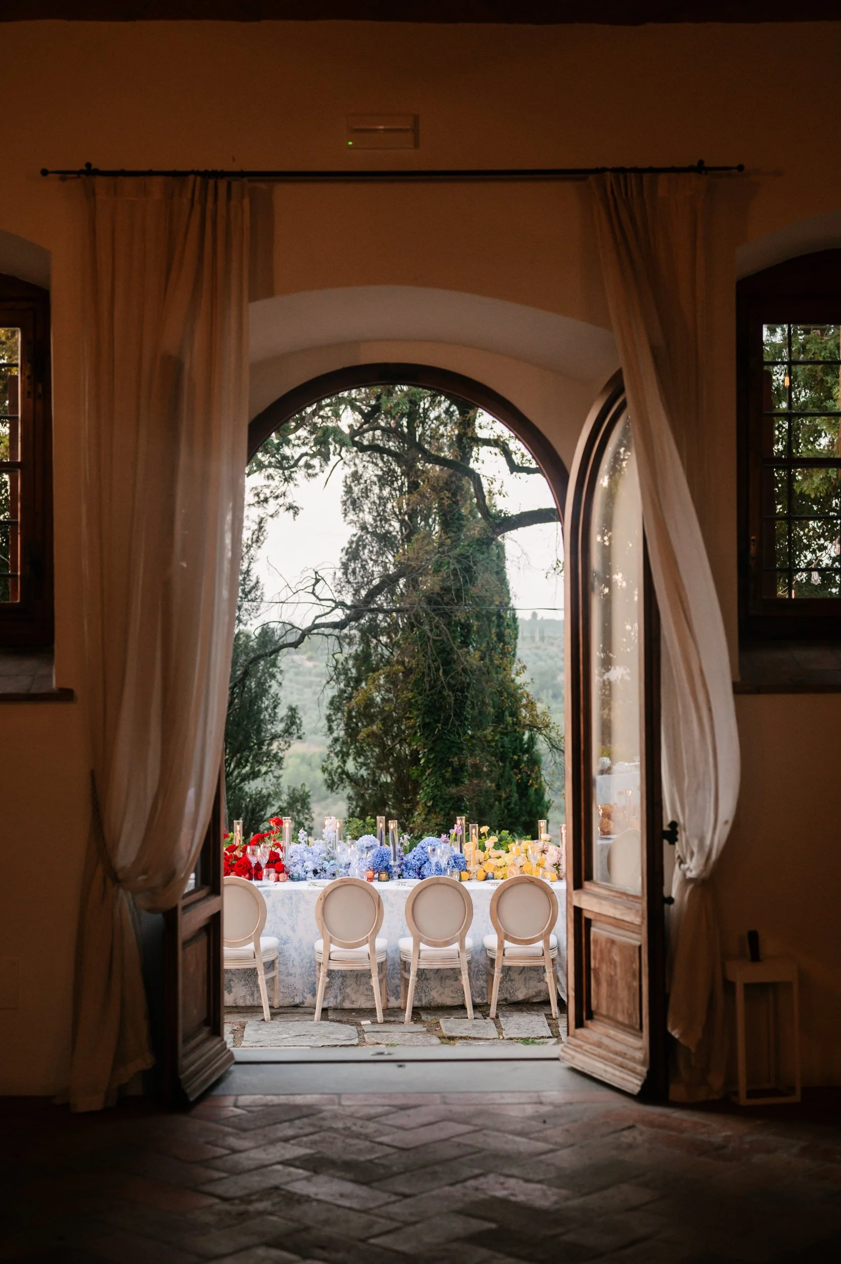 Open door leading to a table set for a celebration outdoors, with flowers and tall candles, in a garden setting with large trees.