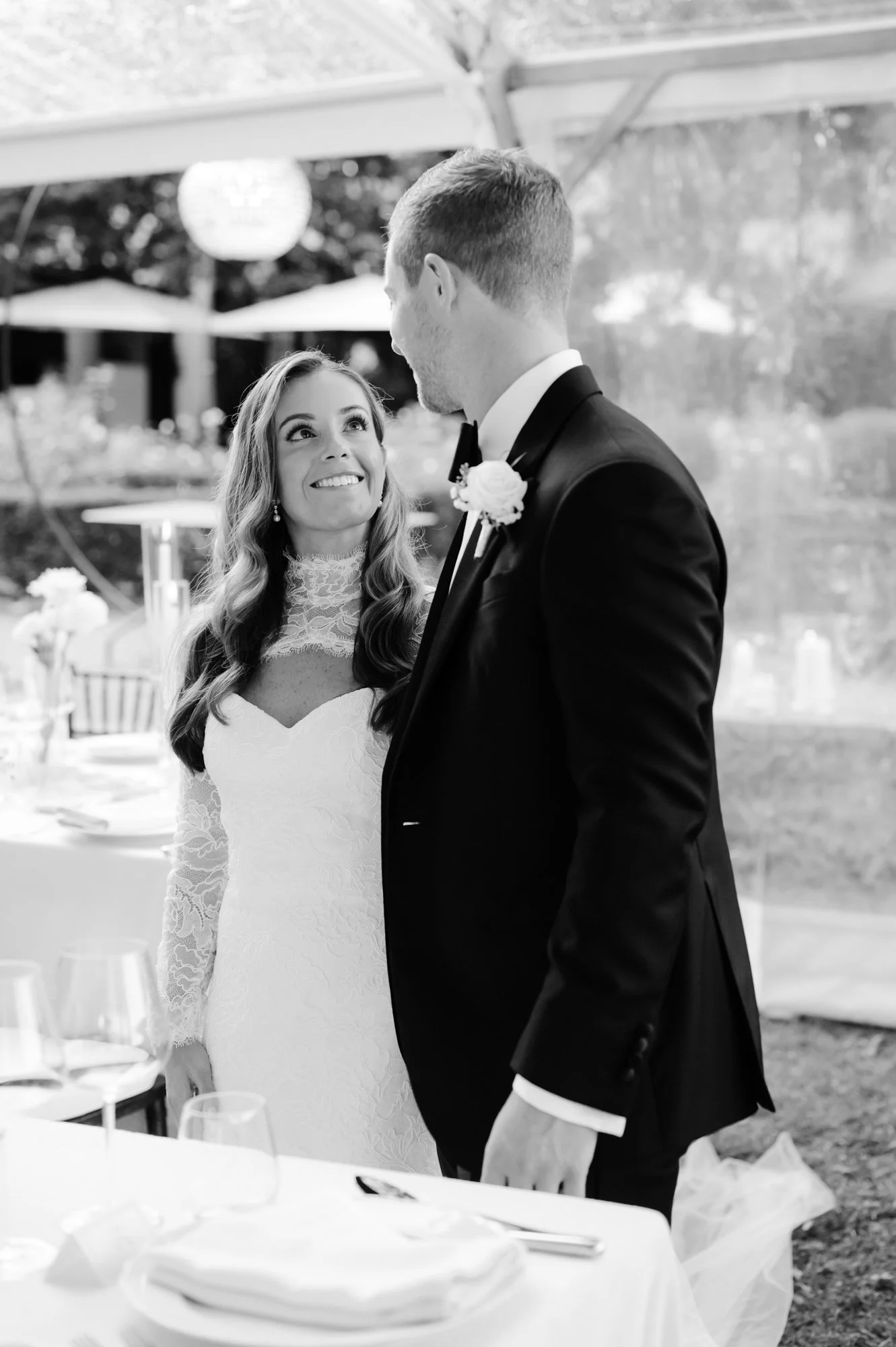 Bride and groom at a wedding reception, standing inside a tent with outdoor scenery visible behind them. The bride has long hair, a lace wedding dress, and is smiling at the groom. The groom is wearing a tuxedo and looking at the bride.