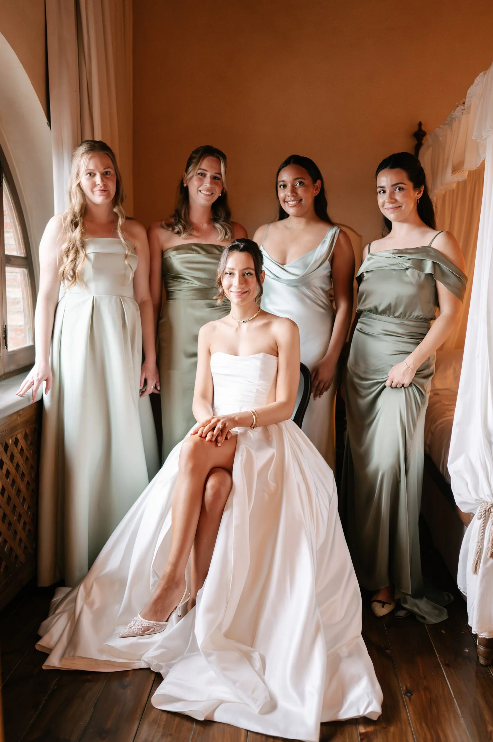 A bride in a white wedding dress sitting on a chair with five bridesmaids in satin dresses standing behind her in a warmly lit room.