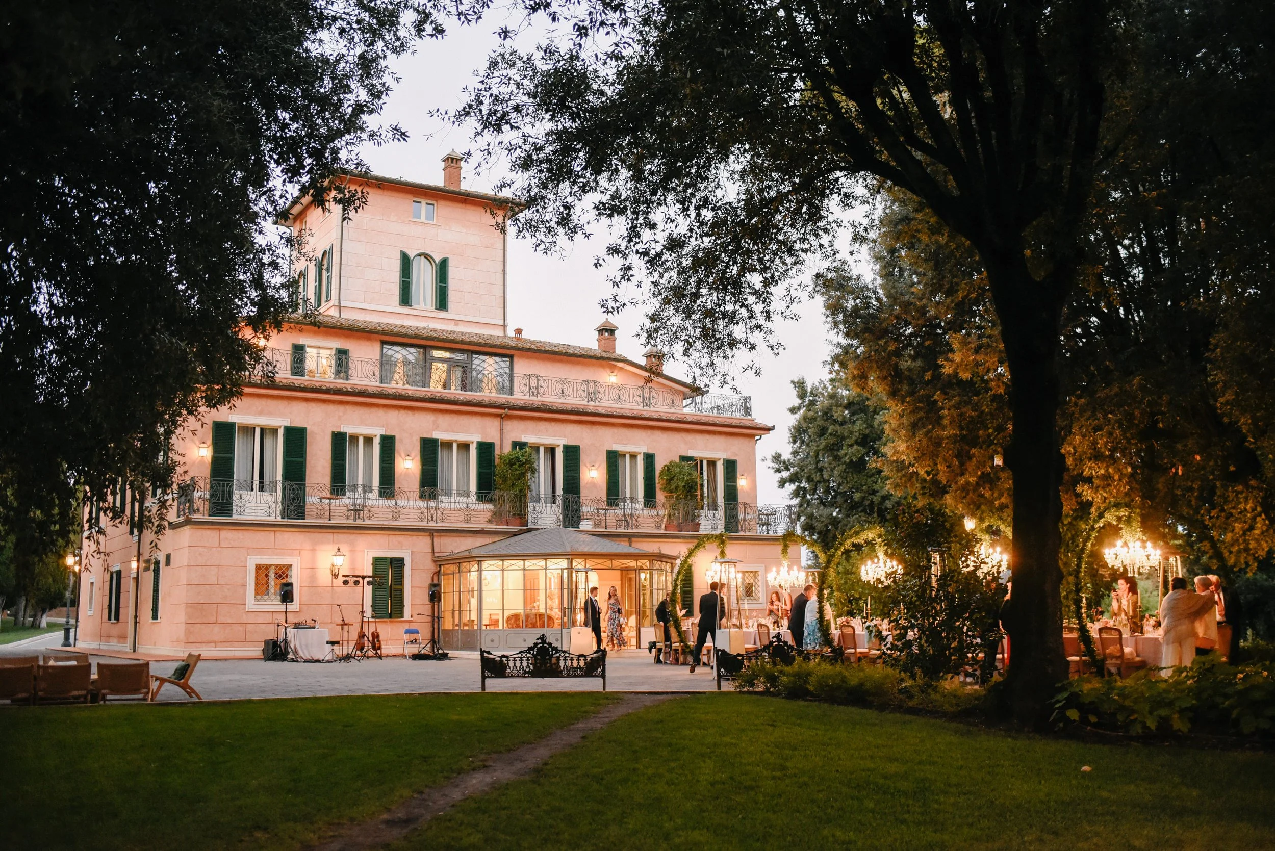 Large pink villa with green shutters and multiple balconies, set amidst trees, illuminated for evening event with guests, tables, and chandeliers outside.