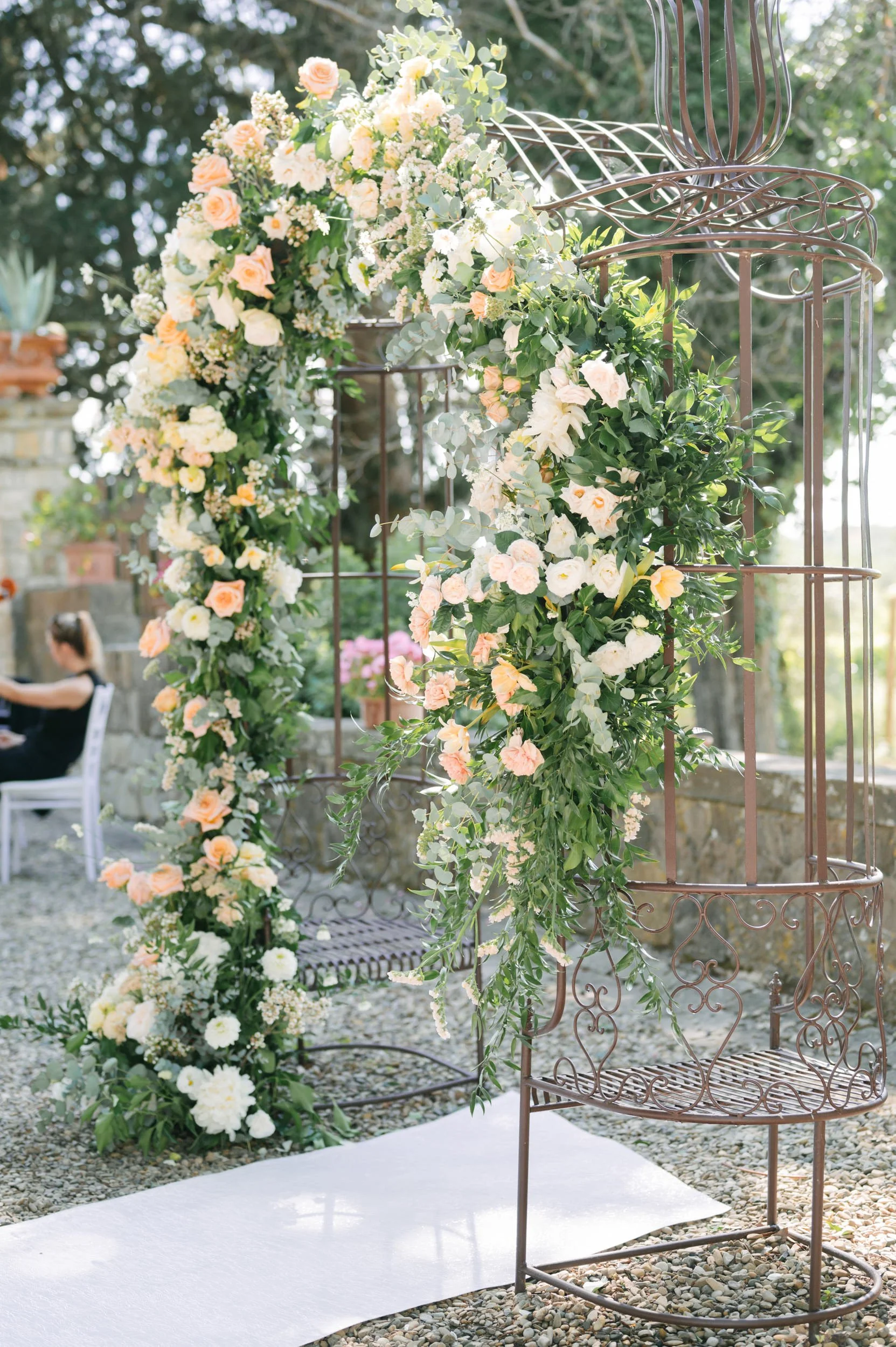 A floral arch decorated with white and blush pink flowers and green foliage, set outdoors with a white aisle runner on gravel ground and a vintage-style chair nearby.