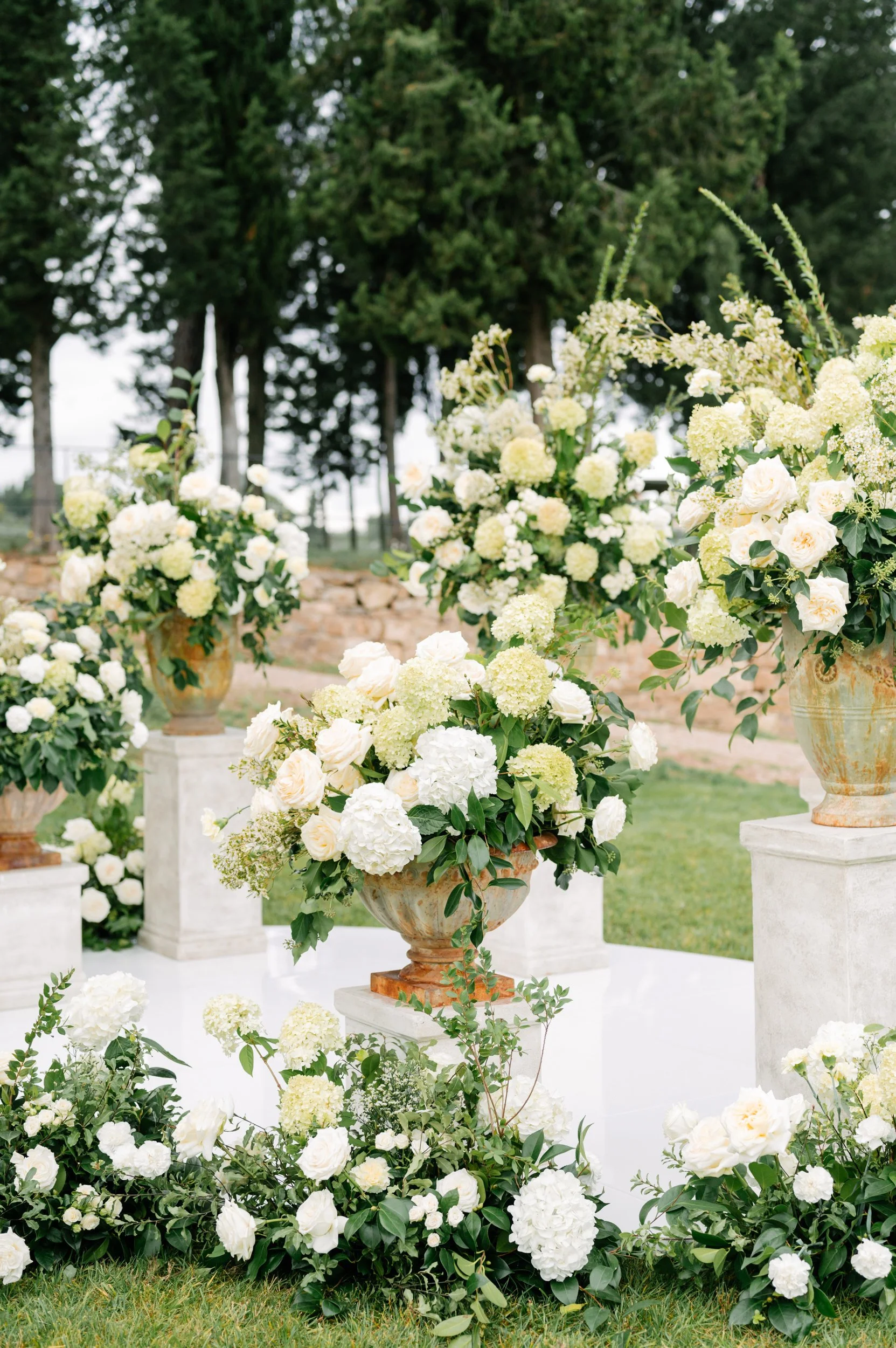 Elegant outdoor floral arrangement featuring large white hydrangeas, roses, and greenery in terracotta and stone vases during daytime.