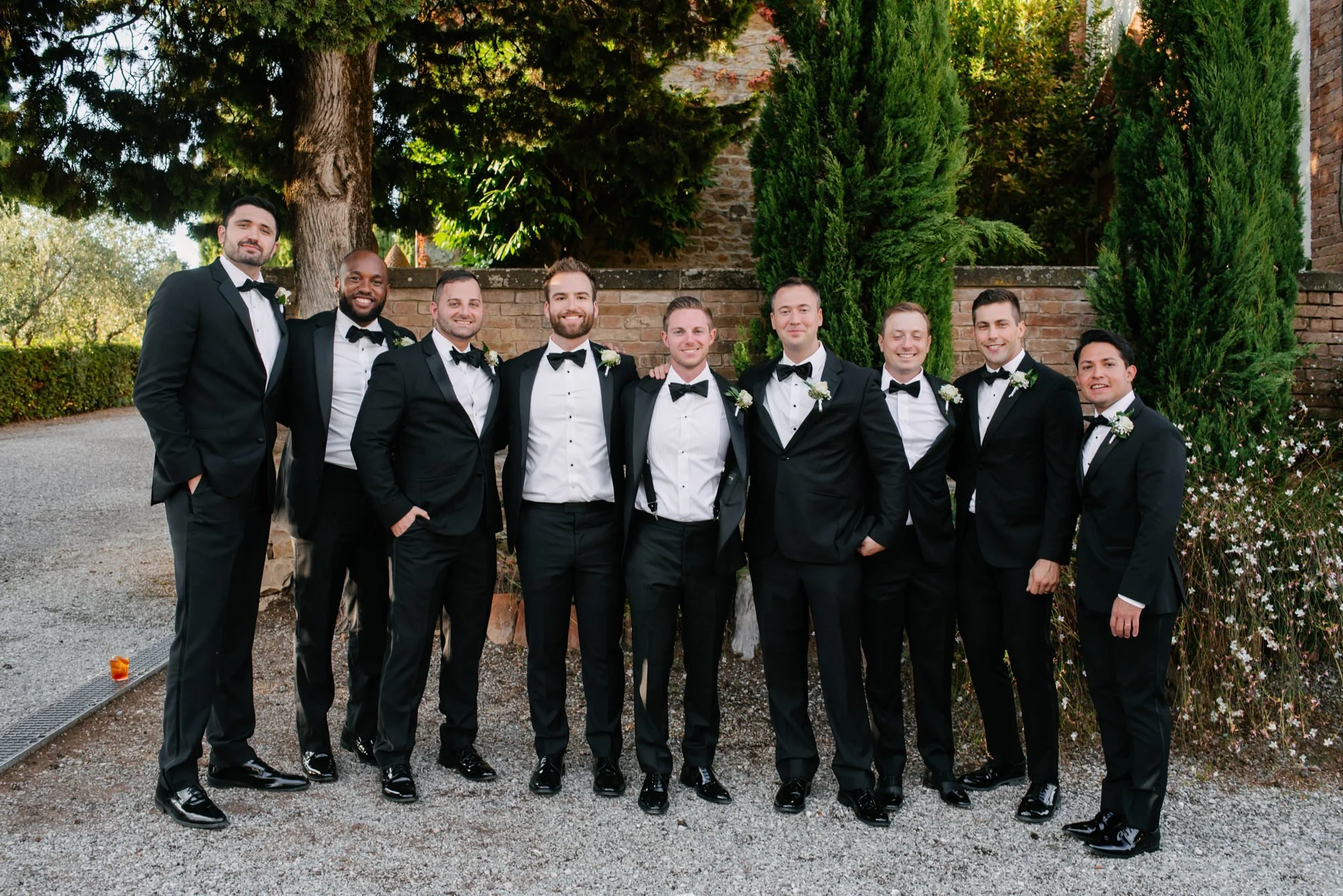Group of ten men in tuxedos standing outdoors in front of trees and a brick wall, smiling for a photo during a formal event.