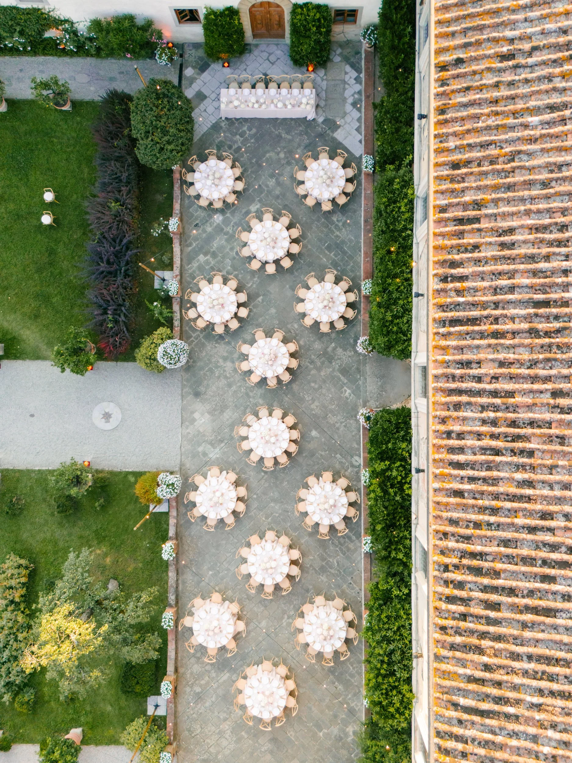 Overhead view of an outdoor event setup with round tables covered with white tablecloths, surrounded by chairs, on a paved patio area. The area is decorated with greenery, flowers, and small lights along the edges, adjacent to a building with a tiled