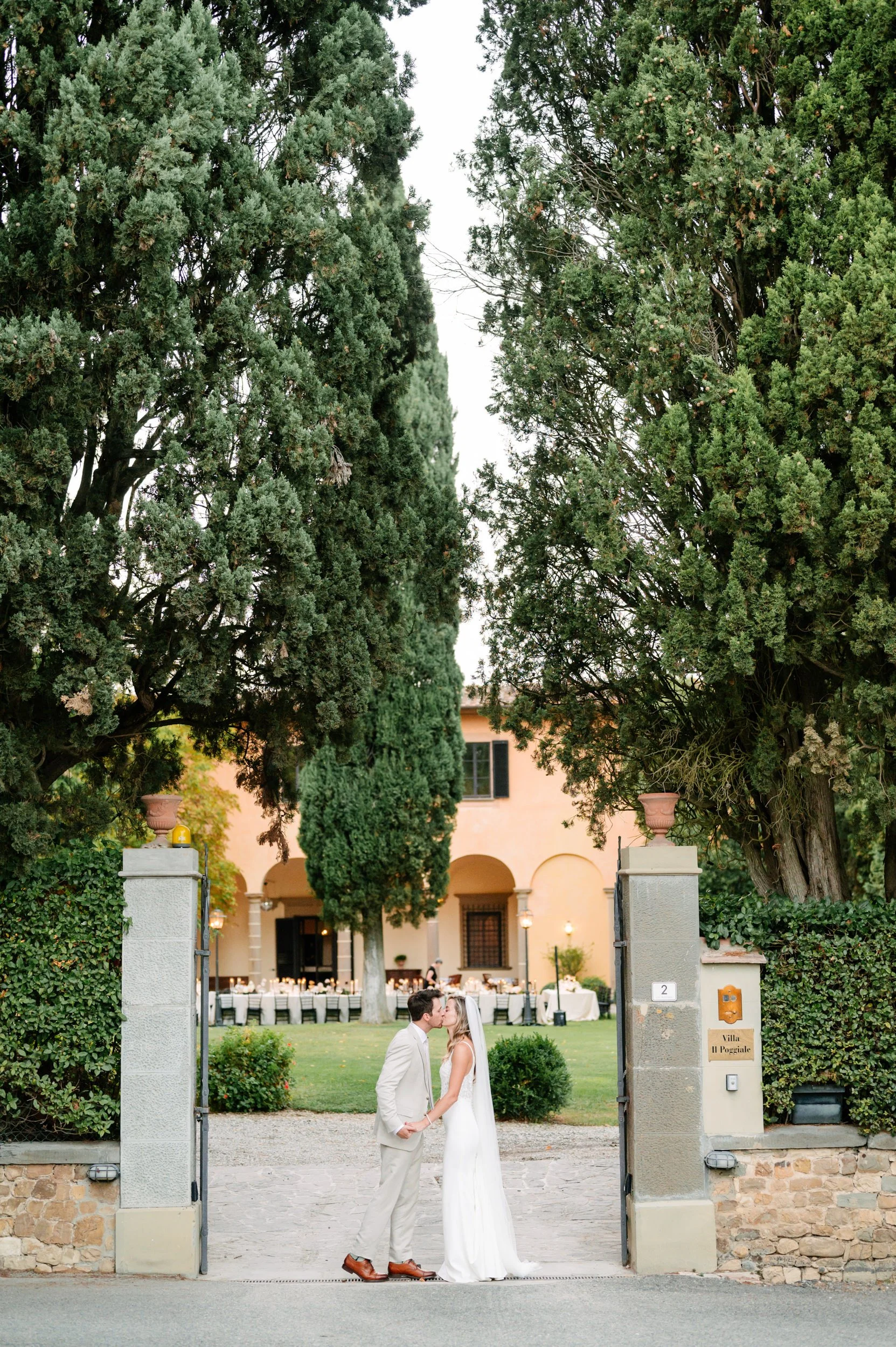 A bride and groom kissing outside a gated estate with lush greenery and a pink villa in the background