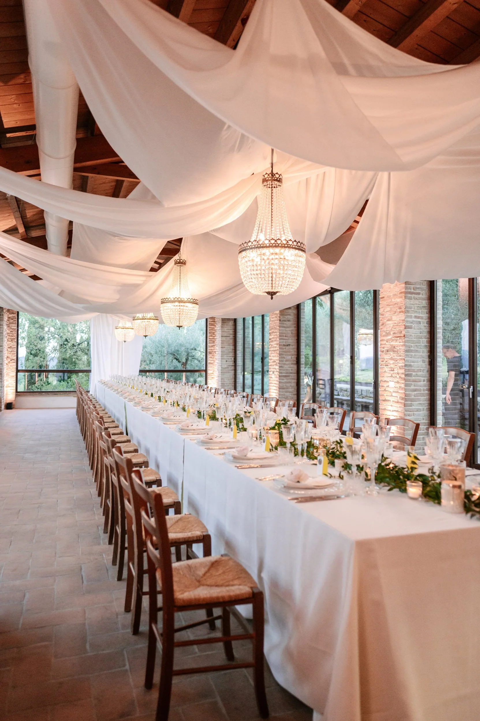 Elegant banquet hall decorated for a wedding reception with long white table, napkins, glassware, candles, and greenery, with draped fabric ceiling and chandeliers, large windows, and brick walls.