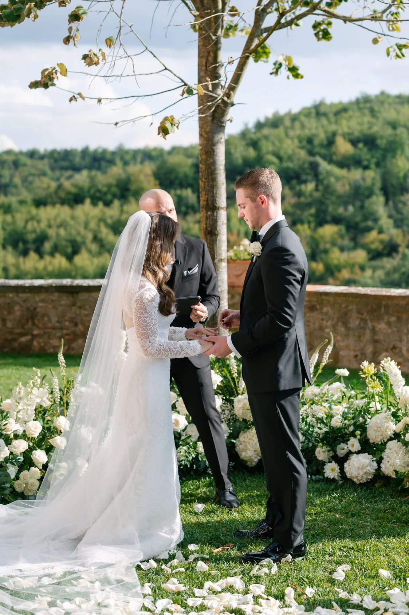 A couple getting married outdoors at a wedding ceremony, with an officiant and floral arrangements, under a tree with a scenic green landscape in the background.