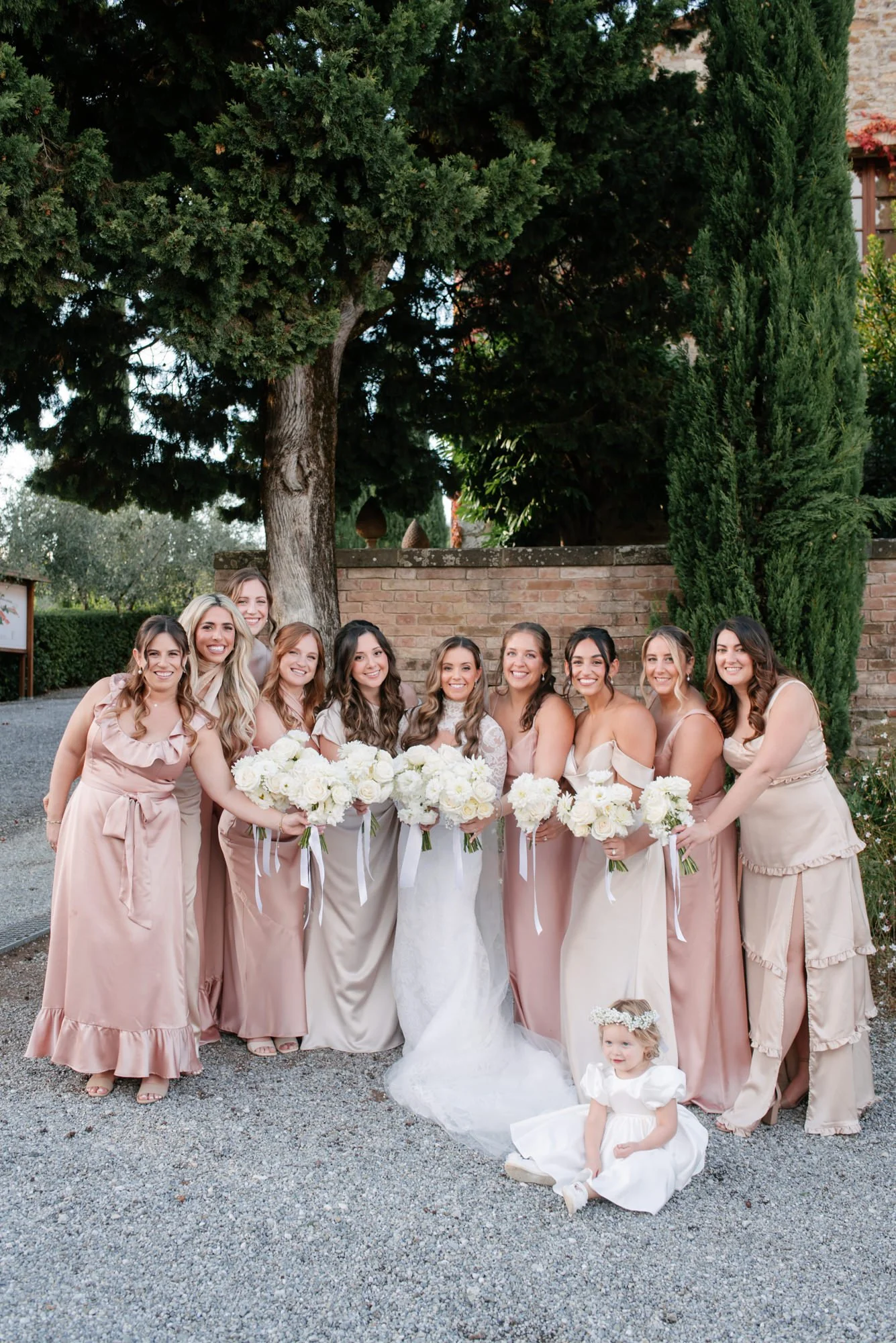 A group of women in light-colored dresses and a young girl in a white dress, holding bouquets of white flowers, posing outdoors in front of a brick wall and large trees during a wedding celebration.