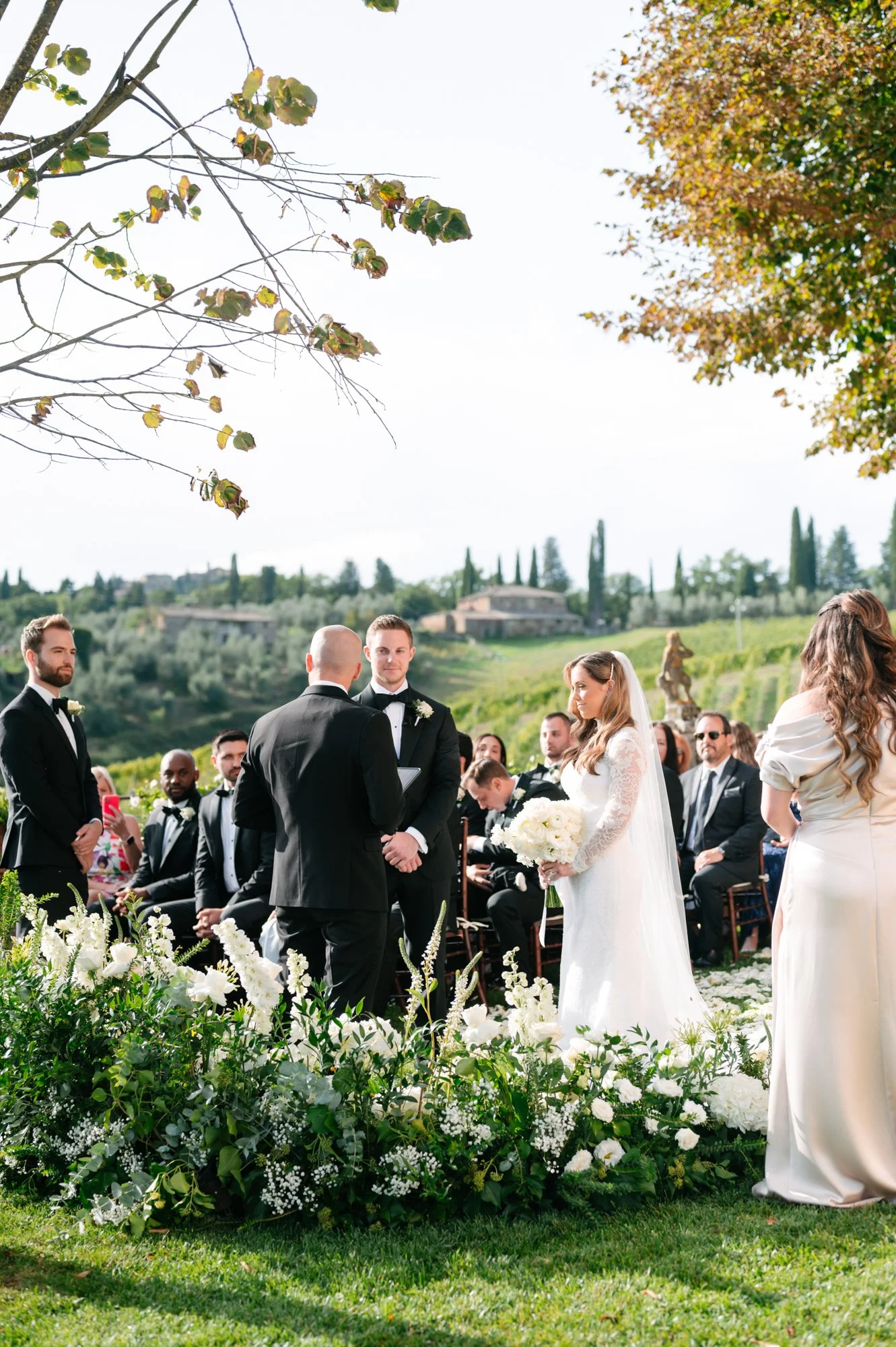 A wedding ceremony outdoors with the bride, groom, and officiant standing in front of seated guests. The bride is holding a bouquet, and the ceremony is set amidst greenery with a scenic landscape in the background.