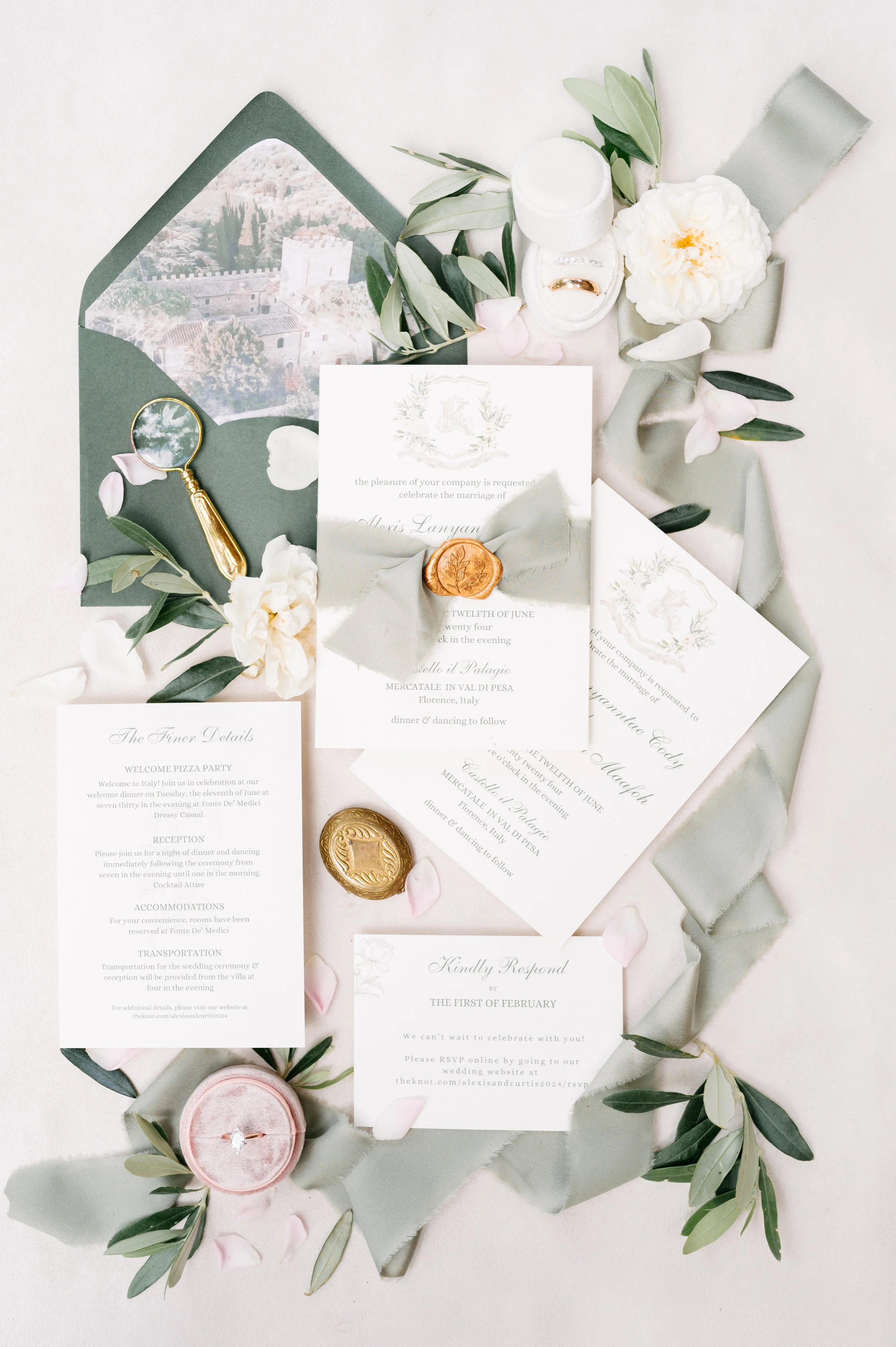 Wedding invitation suite with floral accents, gold wax seals, a small magnifying glass with gold handle, and a ring in a pink velvet box, all arranged on a light surface.