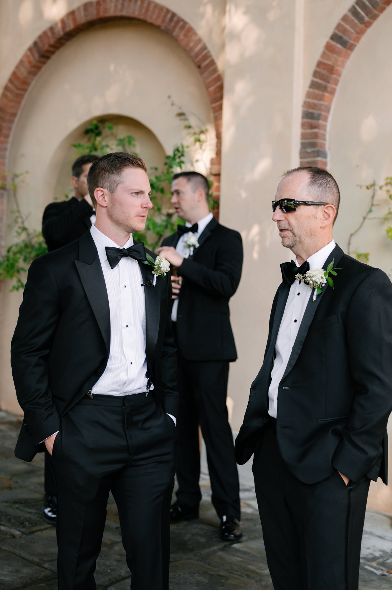 Four men in tuxedos at a formal event, engaged in conversation outside near an archway with decorative brick and greenery.