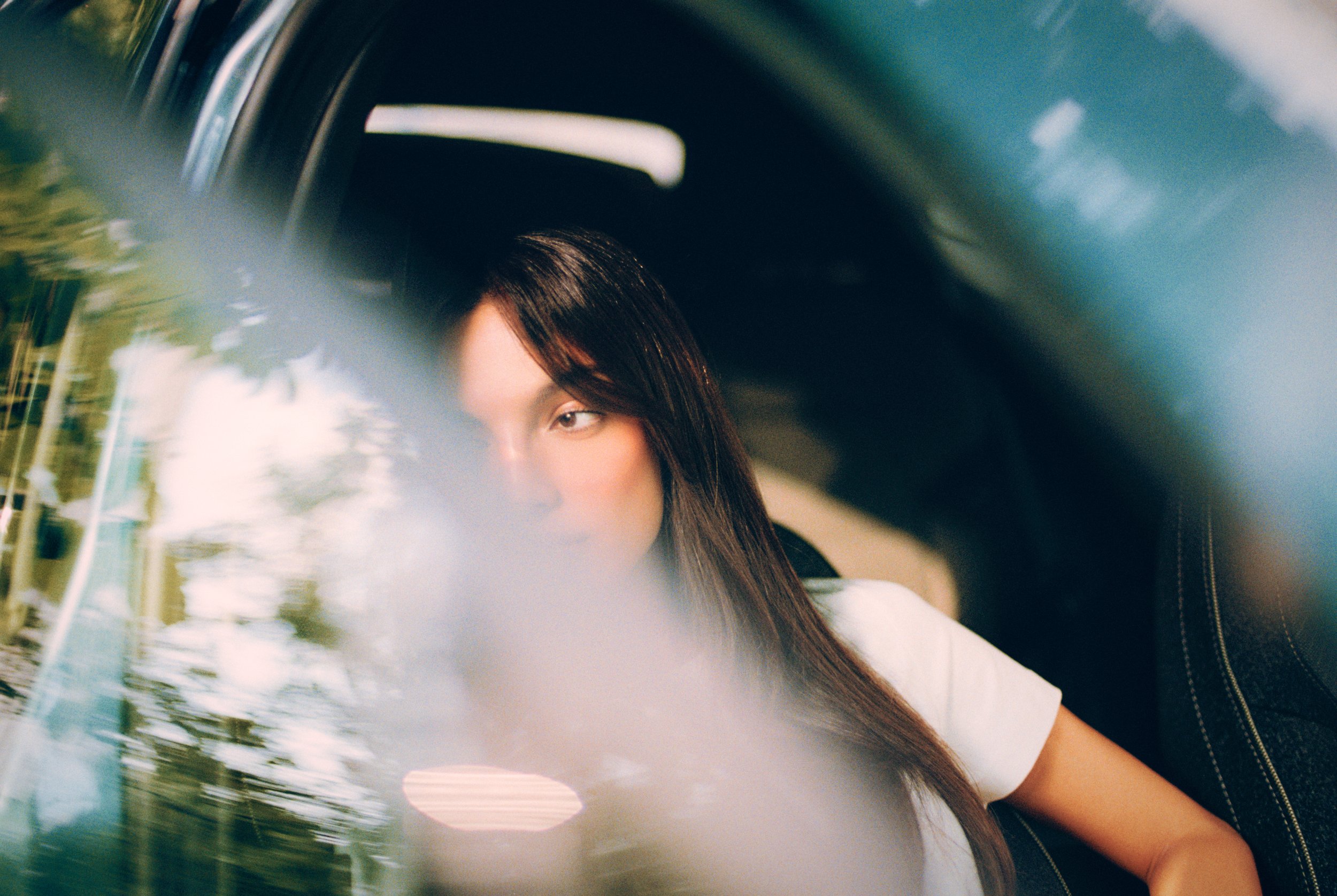 A person with long dark hair and light skin sitting inside a vehicle, viewed through a reflective surface showing trees and sky outside.