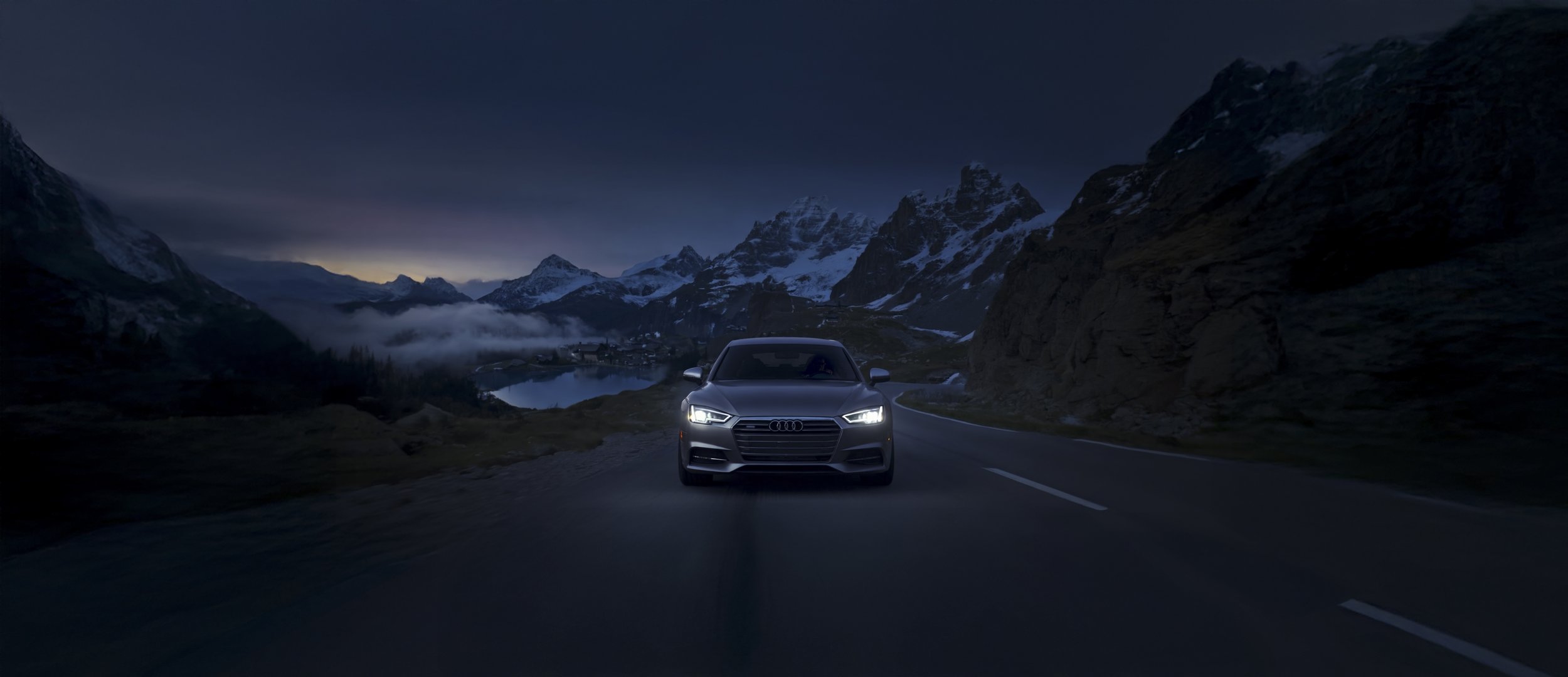 A silver Audi car driving on a mountain road at dusk, surrounded by snow-capped mountains and a lake in the background.