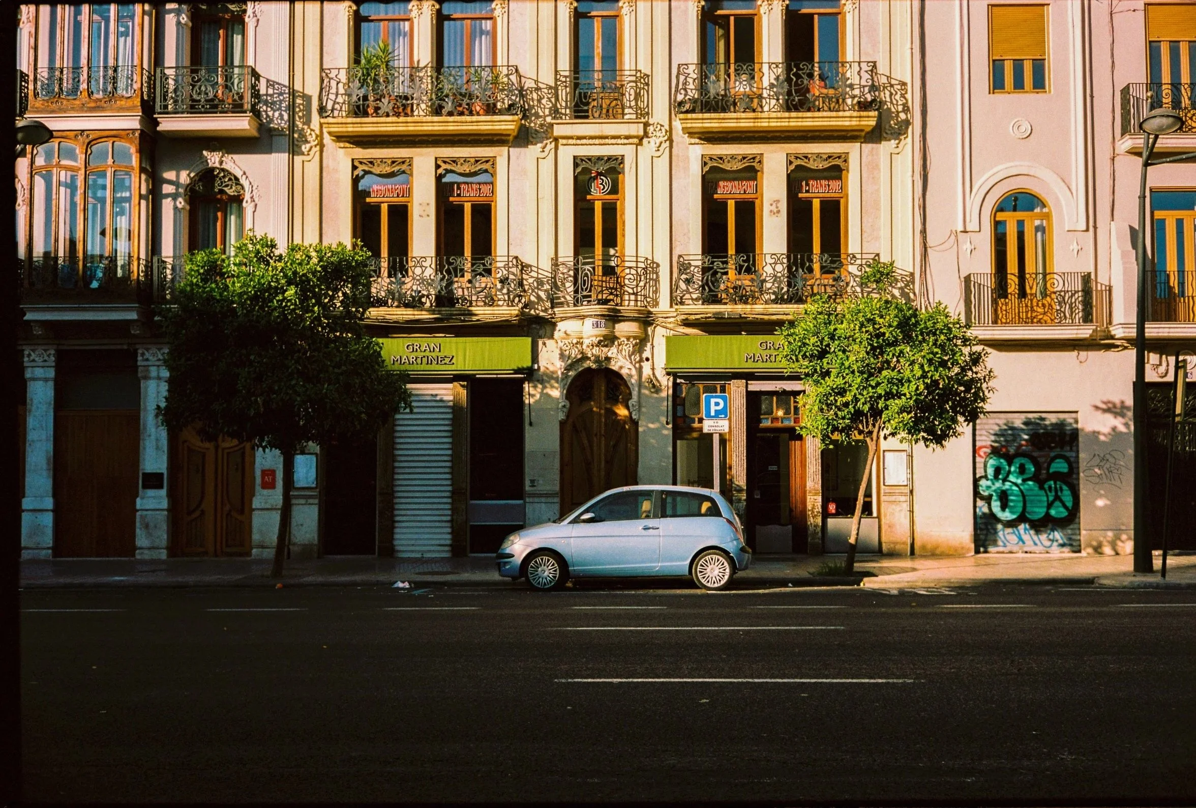 A city street scene with a silver car parked in front of colorful apartment buildings with ornate balconies, trees, and streetlights.