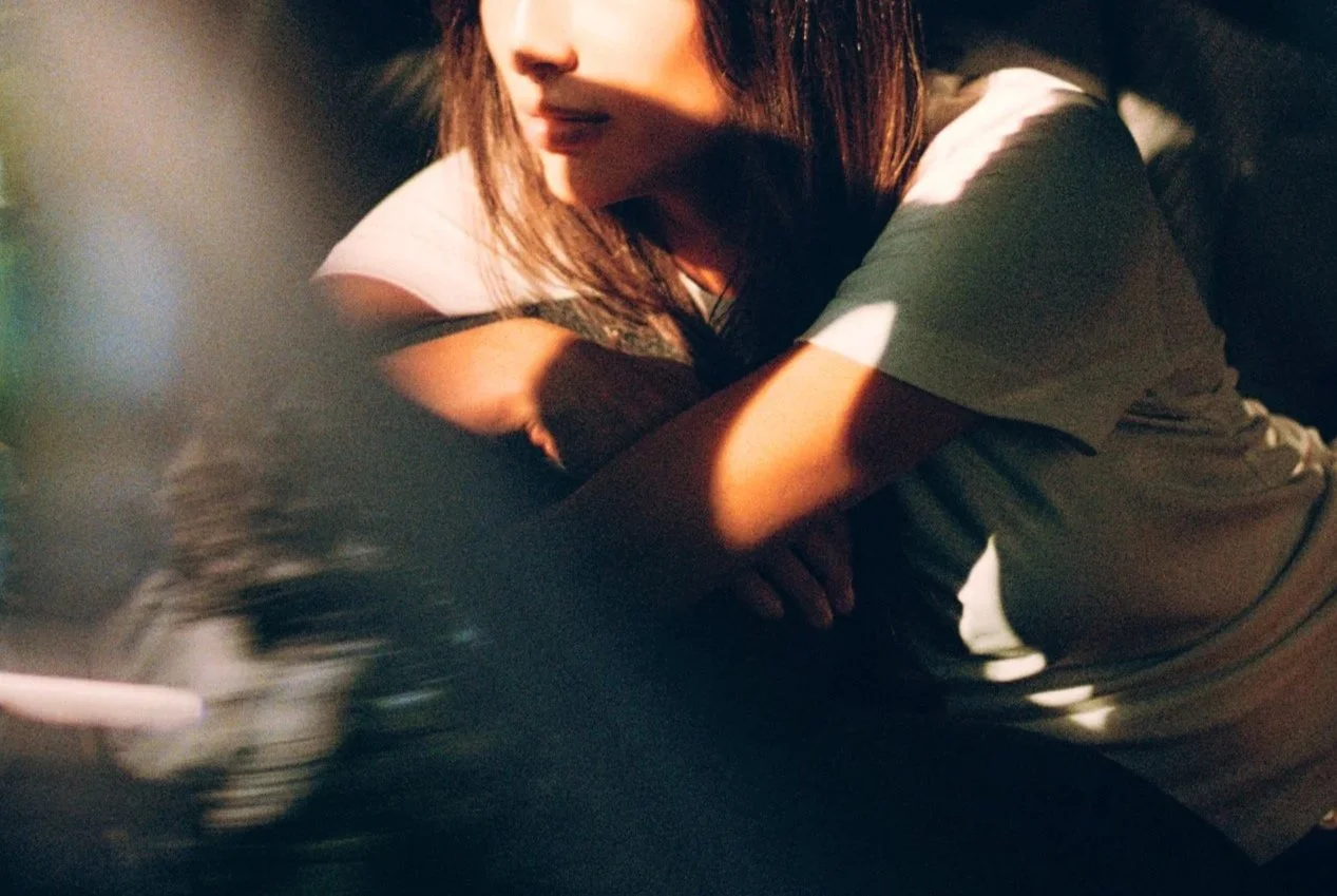 A woman with dark hair is resting her head on her arms, with shadows falling across her face and left arm, in a dimly lit environment.