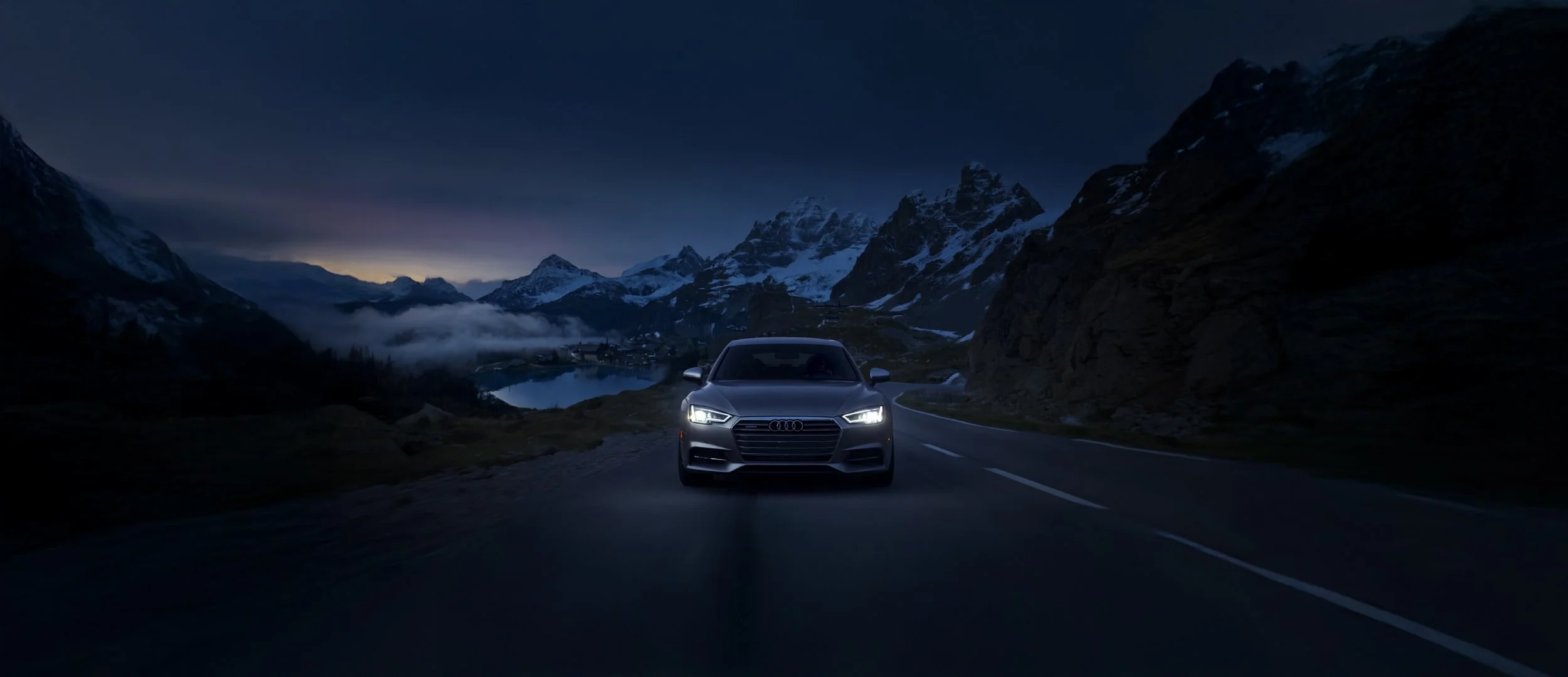 A silver Audi car driving on a mountain road at night with snow-capped peaks and a lake in the background.