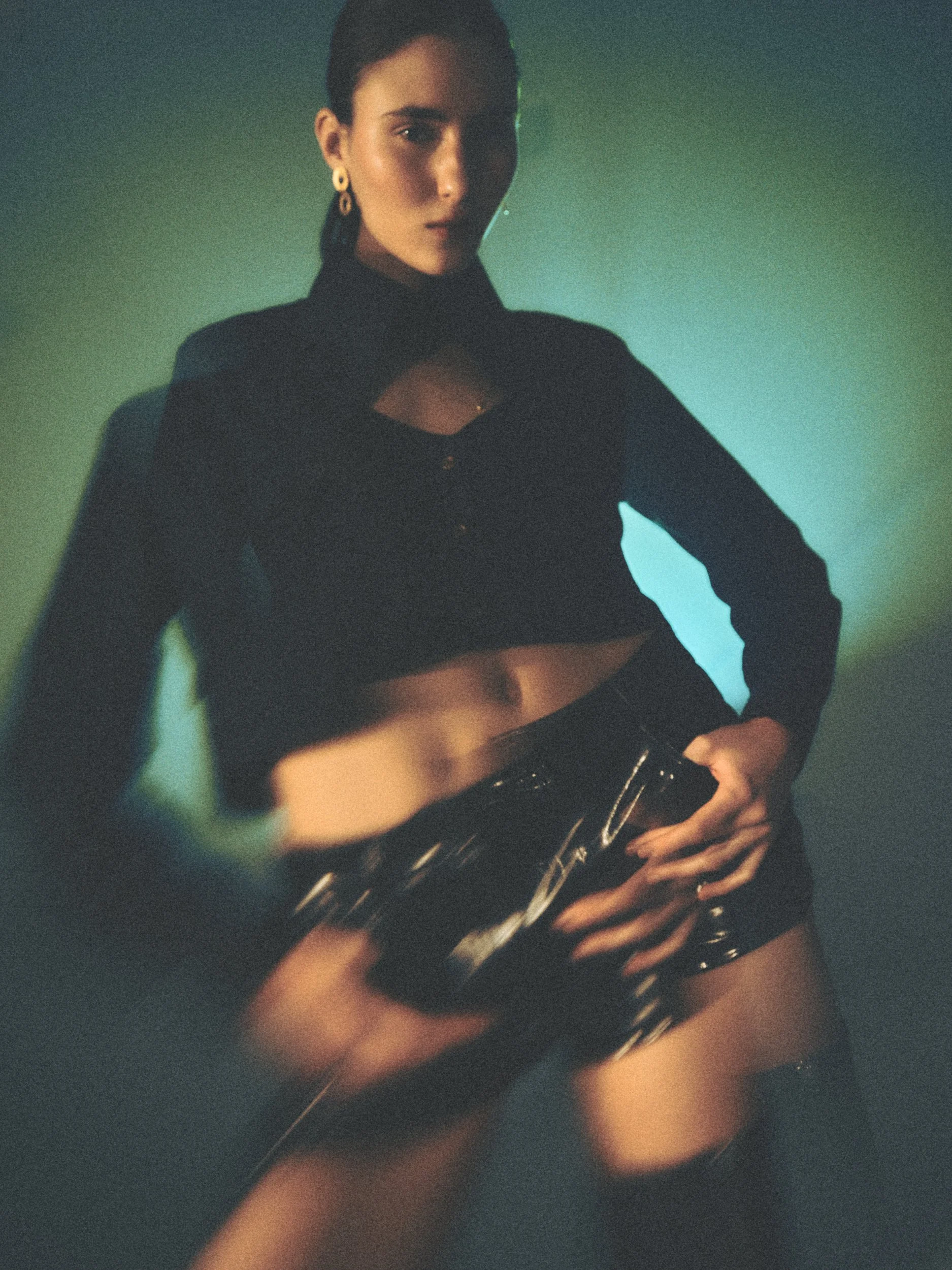 A woman dressed in dark clothing, with one hand on her hip, wearing gold earrings, and standing against a gradient background.