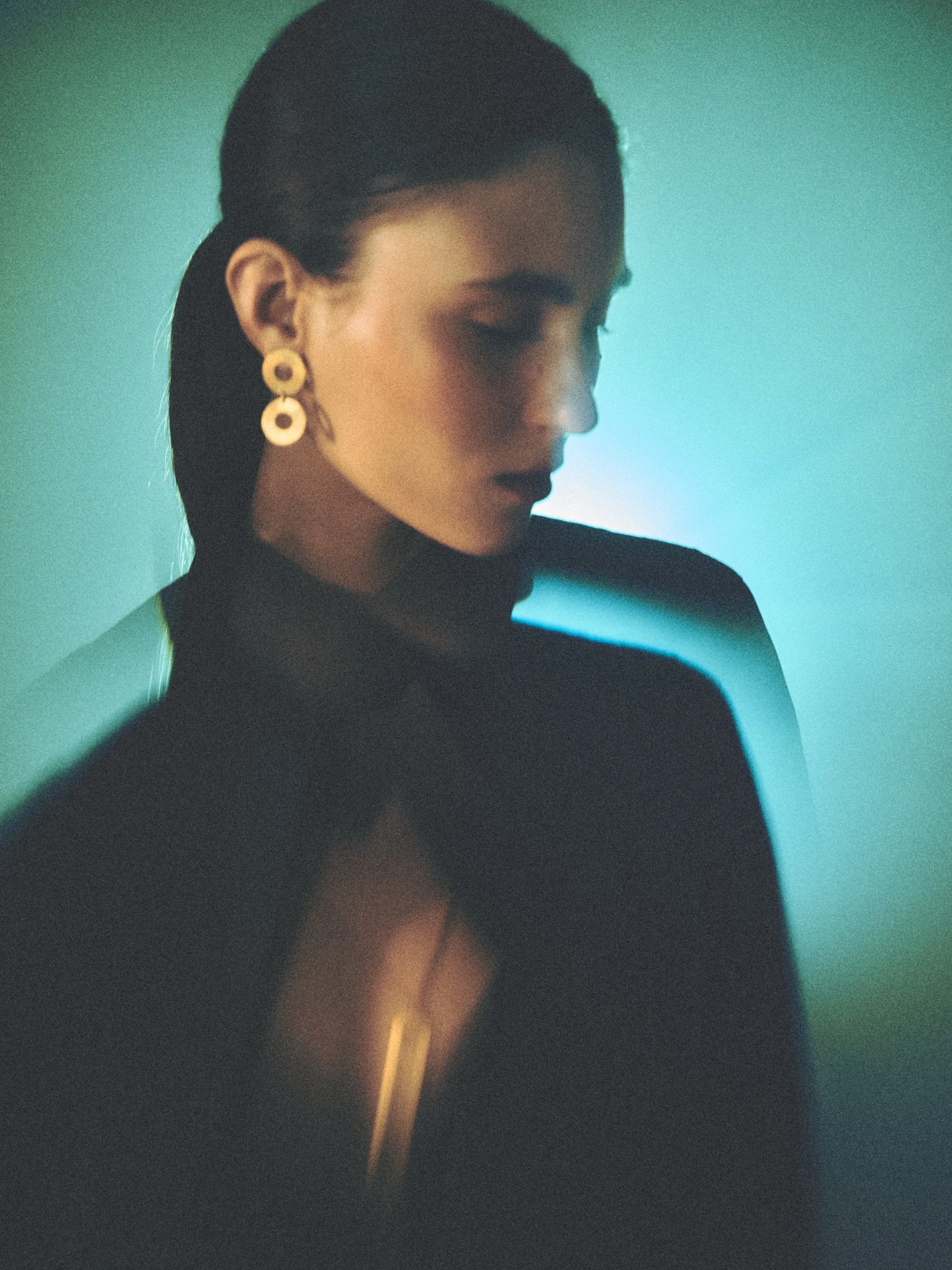 A woman with dark hair tied back, wearing large gold earrings and a black top, is looking down with her eyes closed. The background features a gradient of blue and green hues, and a golden vertical streak is present in the image.