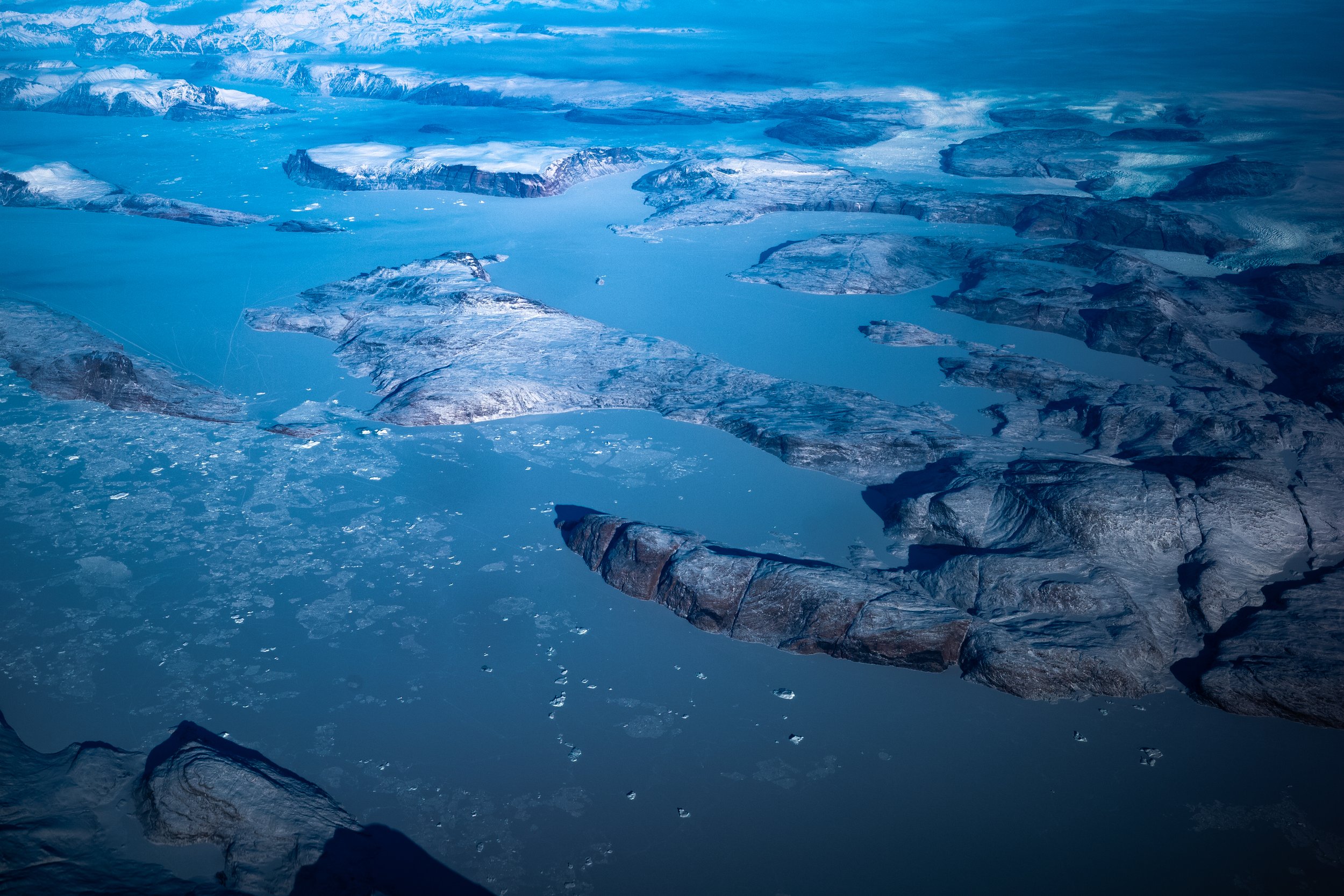 Aerial view of ice-covered landscape with a frozen lake surrounded by snow-covered mountains and rocky terrain in the background.
