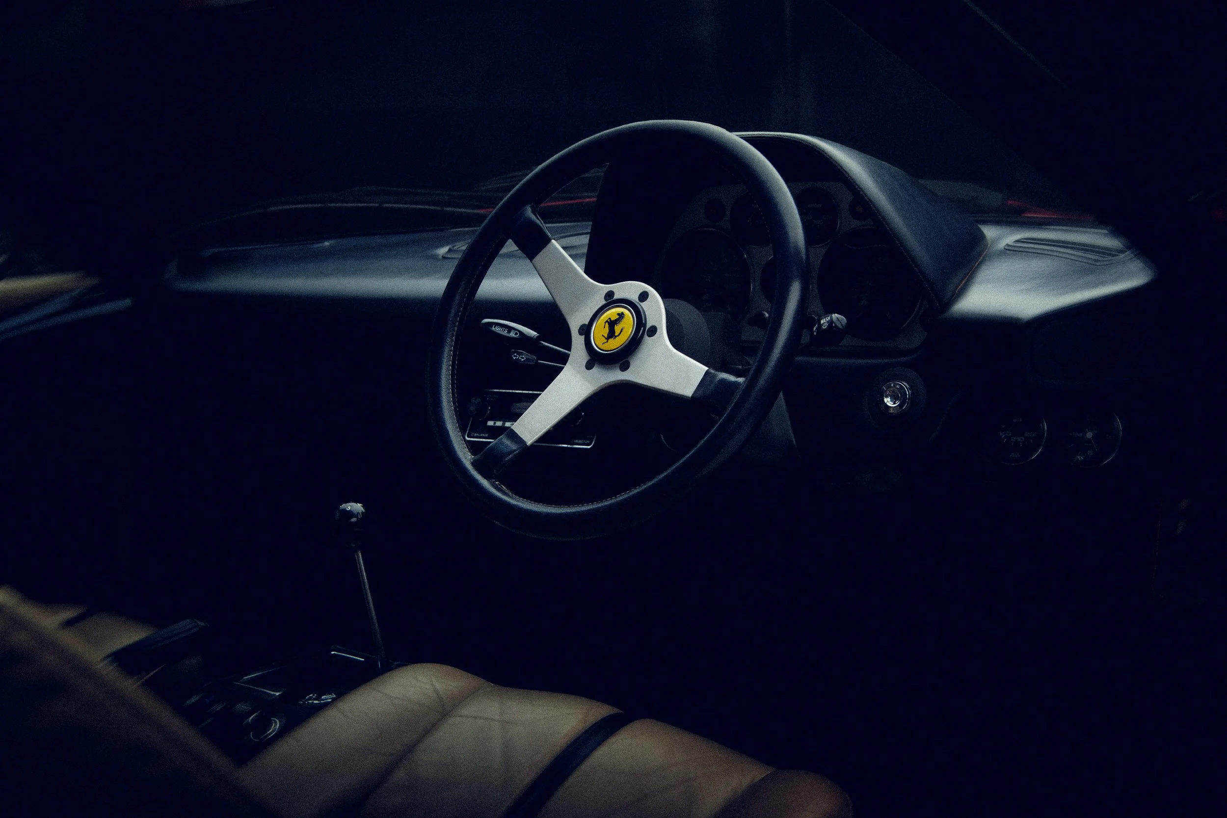 The interior of a classic Ferrari car, showing a steering wheel with the Ferrari logo, dashboard, and a beige seat.